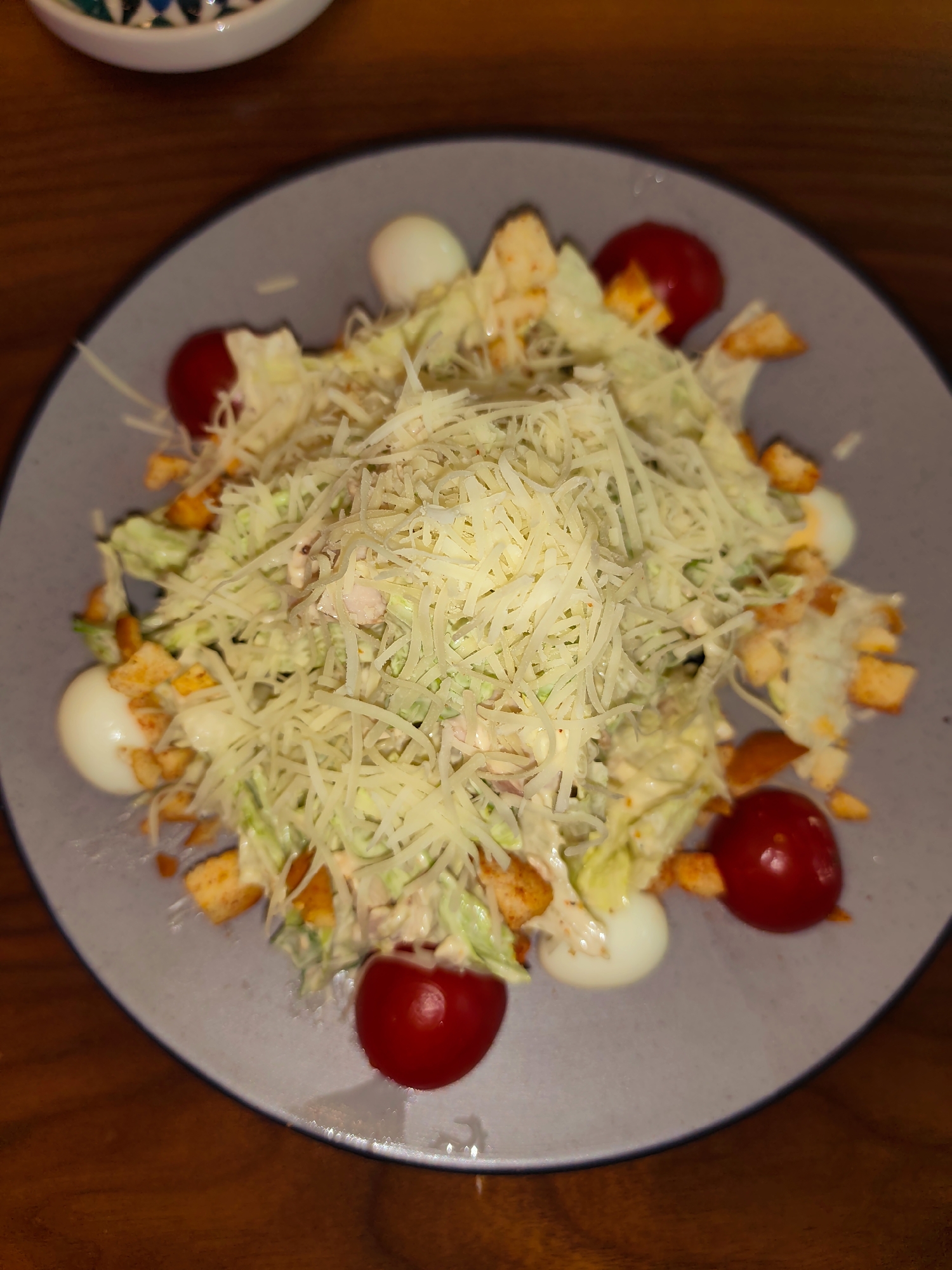 A Caesar salad is presented on a grey plate, which rests on a wooden table. The salad is topped with shredded cheese, croutons, and garnished with cherry tomatoes and boiled quail eggs. The image is taken from a top-down perspective.