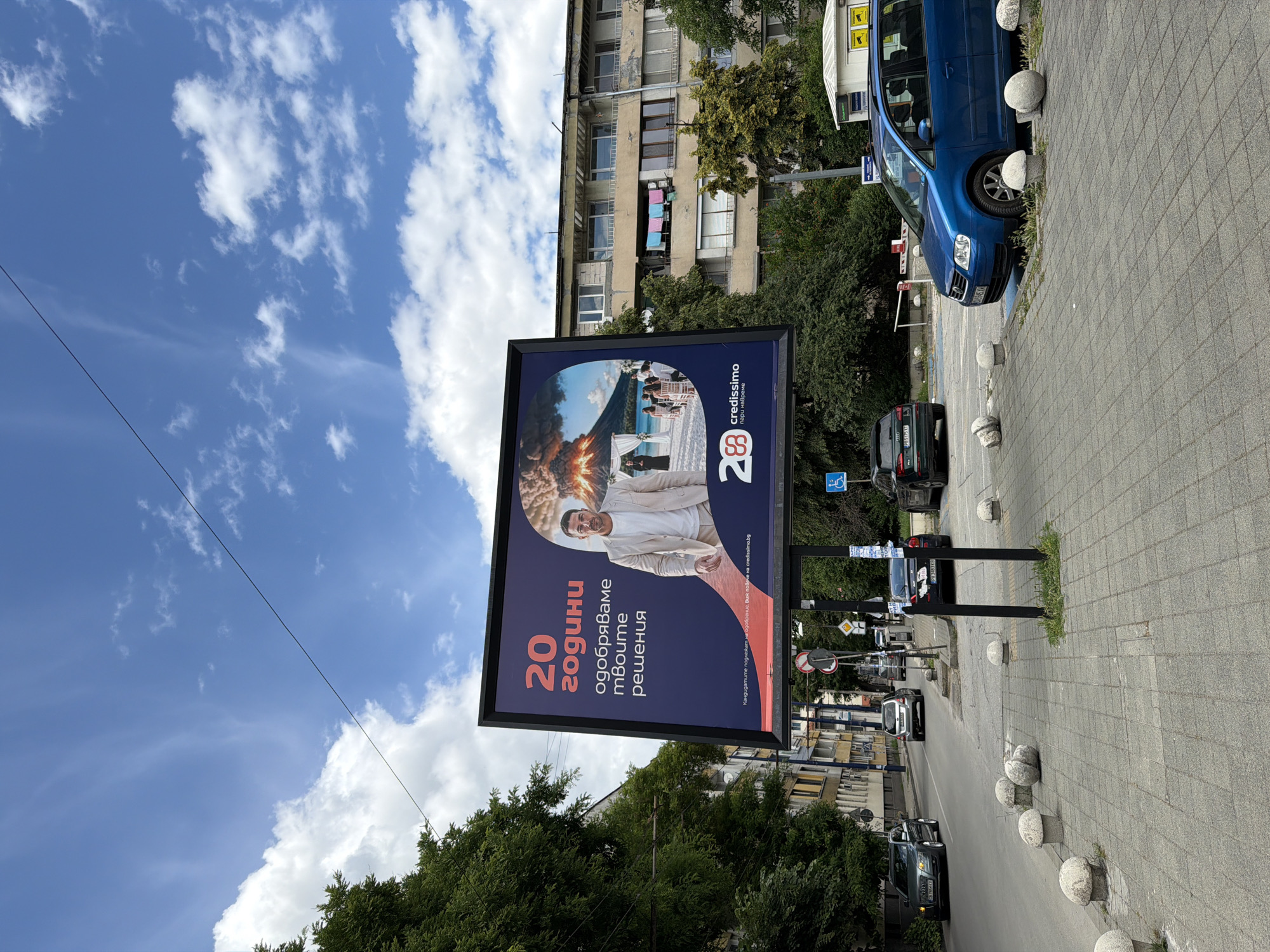 The image captures a billboard in Pazardzhik, Bulgaria, on a bright, cloudy day. The billboard features a man in a light-colored suit, a volcano erupting in the background, and text in Bulgarian. Below the main image, the logo for "credissimo" is visible. The billboard is positioned on a sidewalk next to a street with several cars parked and driving by. In the background, residential buildings and trees are visible. The street is paved with asphalt, and the sidewalk is made of grey tiles. Several spherical concrete bollards are placed along the sidewalk. A blue minivan is parked in front of the billboard.