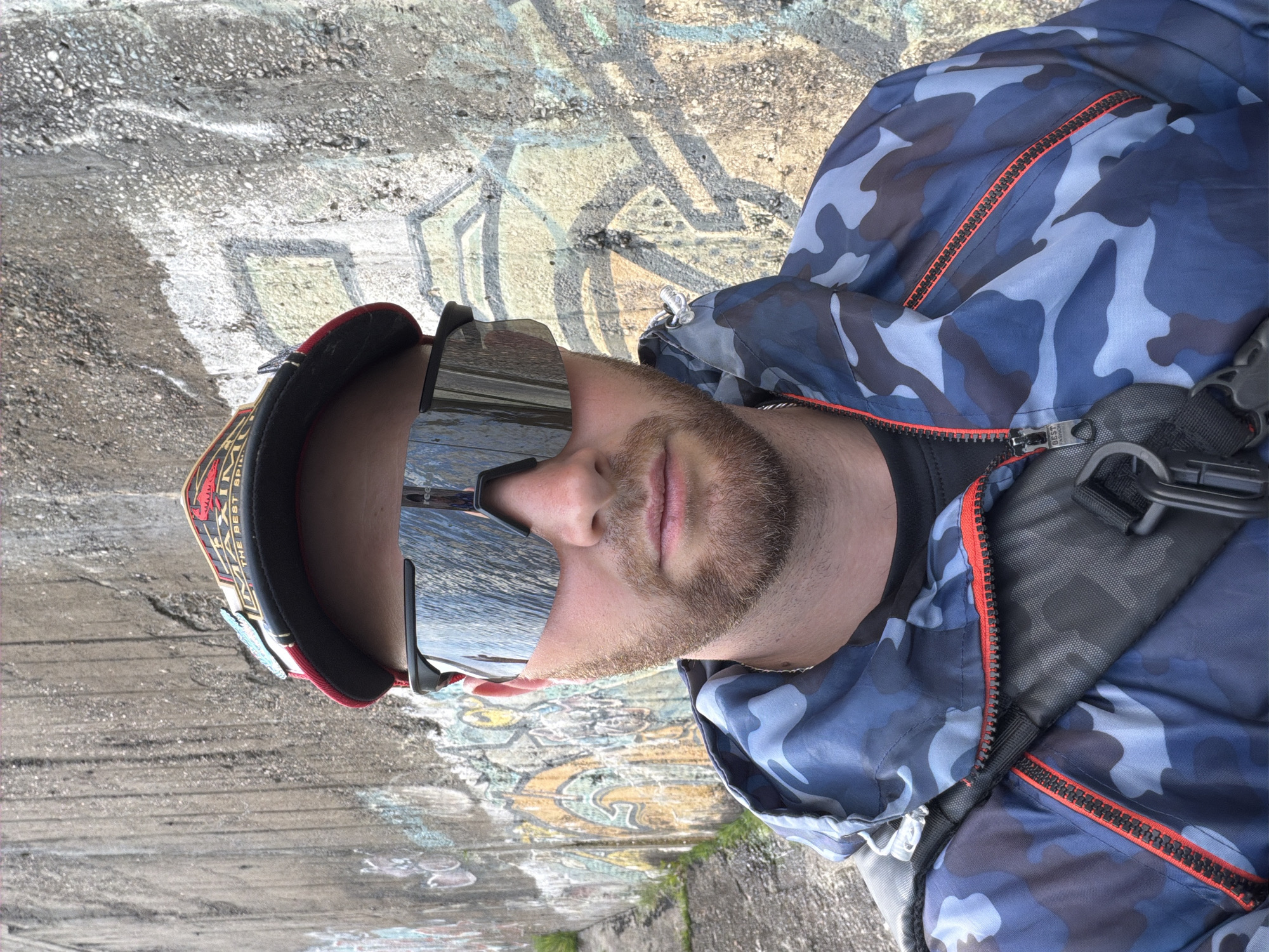 A man is standing outdoors in Chaikovsky, Russia, near a concrete surface covered with graffiti. He is wearing a blue camouflage jacket with a red zipper, reflective sunglasses, and a baseball cap. The man has a beard. He is wearing a black shirt underneath his jacket. He is standing next to a concrete wall. There is some vegetation growing near the wall. A black strap or harness is visible over his right shoulder.