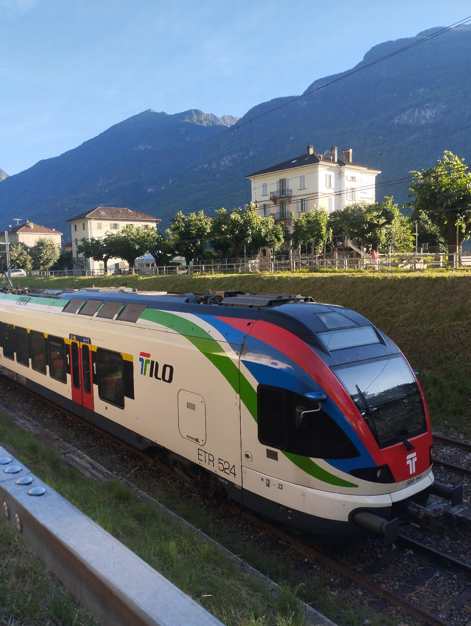 A train with "TRILO" branding and a colorful stripe design is stopped on tracks. The train is white with a red, blue, and green stripe that runs along the top. The front of the train is red and blue. The train has several windows along its side, some of which are dark, and some are tinted to reflect the sky. A "TRILO" logo is visible on the side of the train. The train is numbered "ETR 524". In the background, there are several buildings, trees, and mountains. The buildings are light-colored and appear to be apartments or houses. The trees are green and leafy. The mountains are large and covered in vegetation. The sky is clear and blue, indicating a sunny day. The scene is a common sight in a train station or along a railway line, set against a natural mountainous backdrop. The train appears to be stationary, possibly at a stop.