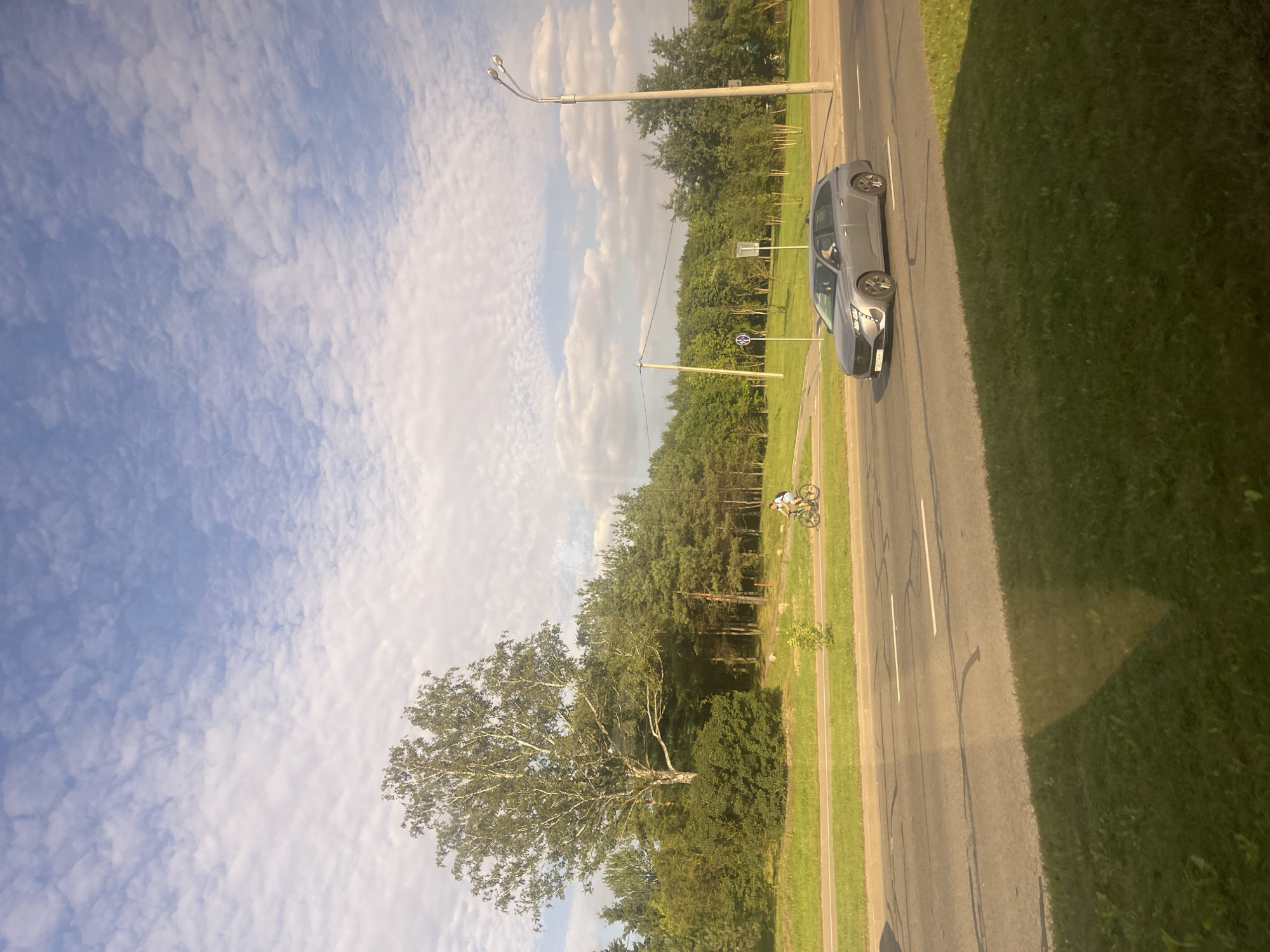 A silver station wagon is driving down a multi-lane road bordered by a green grassy median and a sidewalk with trees. A cyclist is riding on the sidewalk. The sky is blue with scattered white clouds. The scene appears to be in a park or suburban area of Minsk, Belarus.