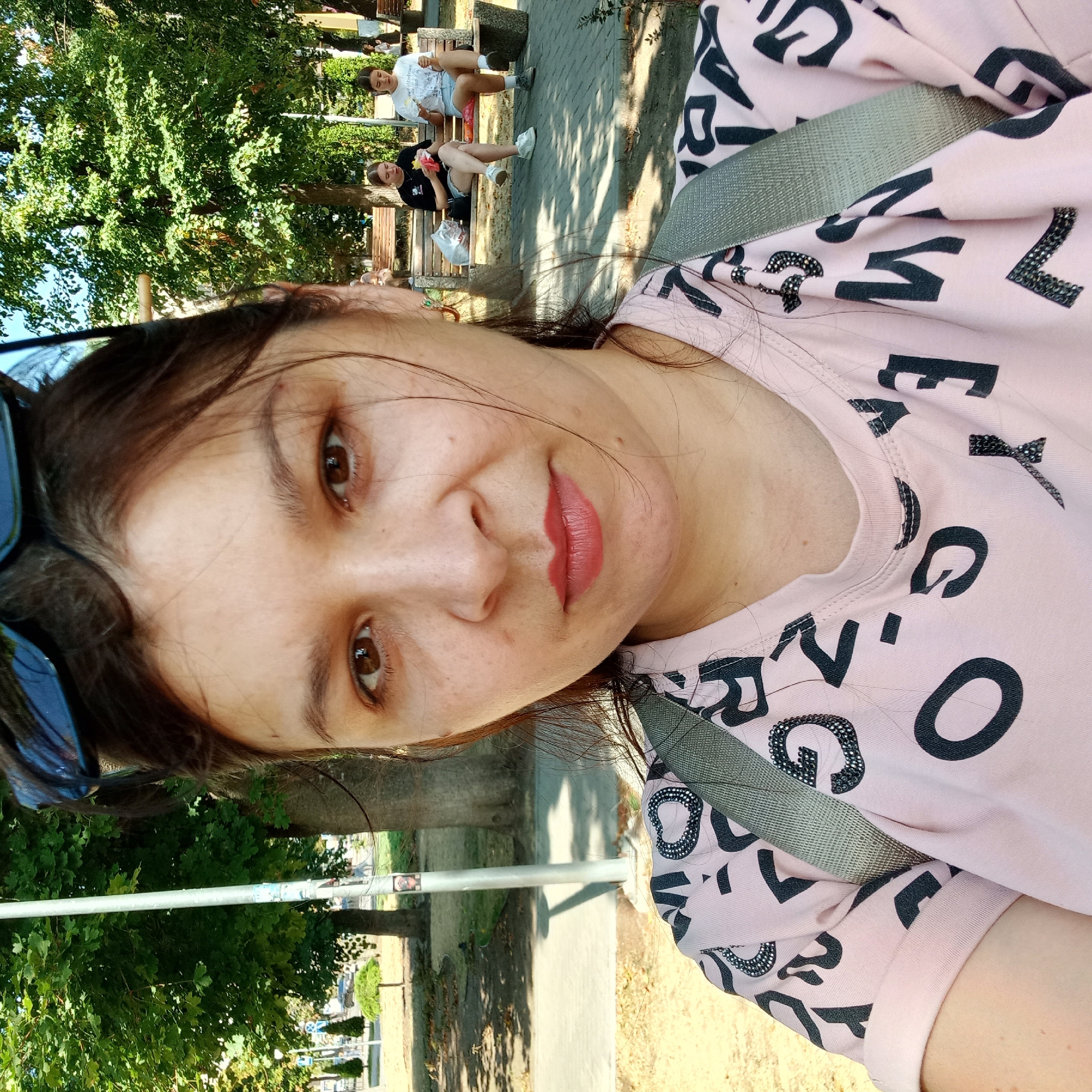 A woman with dark hair and brown eyes is taking a selfie in a park. She is wearing a light pink t-shirt with black letters and a grey backpack strap visible. She has on sunglasses on top of her head and appears to have lipstick on. In the background, two young women are sitting on a park bench eating. The environment is a park with trees, grass, and a paved walkway. The scene appears to be in Kropyvnytskyi, Ukraine, based on the presence of a street lamp with Ukrainian signage.
