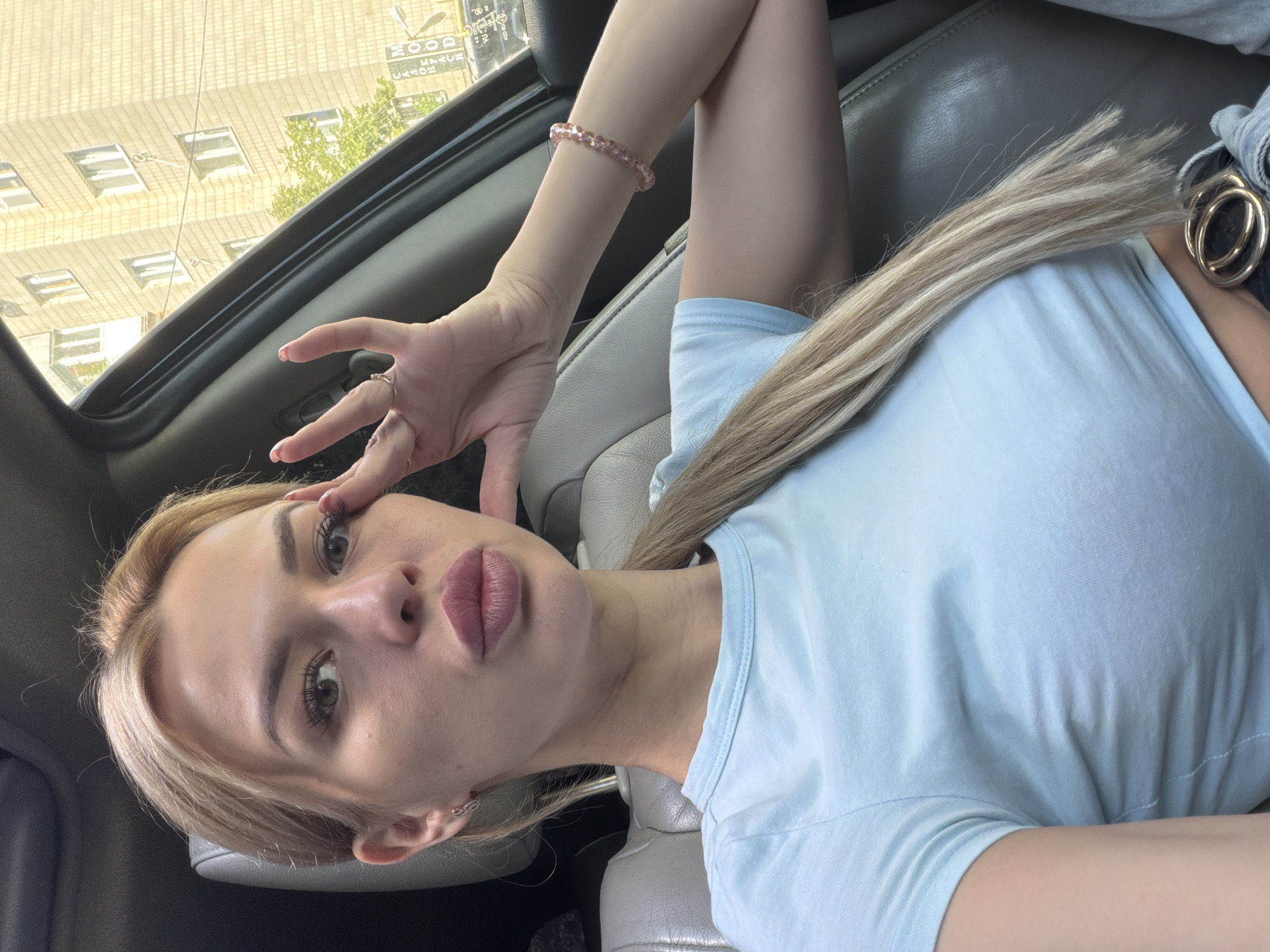 A blonde woman sits in the driver's seat of a car, looking towards the camera. She is wearing a light blue t-shirt. Her right hand is raised to her face, with her fingers touching her eye. She is wearing a ring on her right ring finger and a pink beaded bracelet on her right wrist. Her hair is pulled back into a ponytail. In the background, a multi-story building can be seen, with trees and a street sign. The car interior is visible, with leather seats and a black dashboard. The scene appears to be set in Kyiv, Ukraine, given the context of the building and street sign.