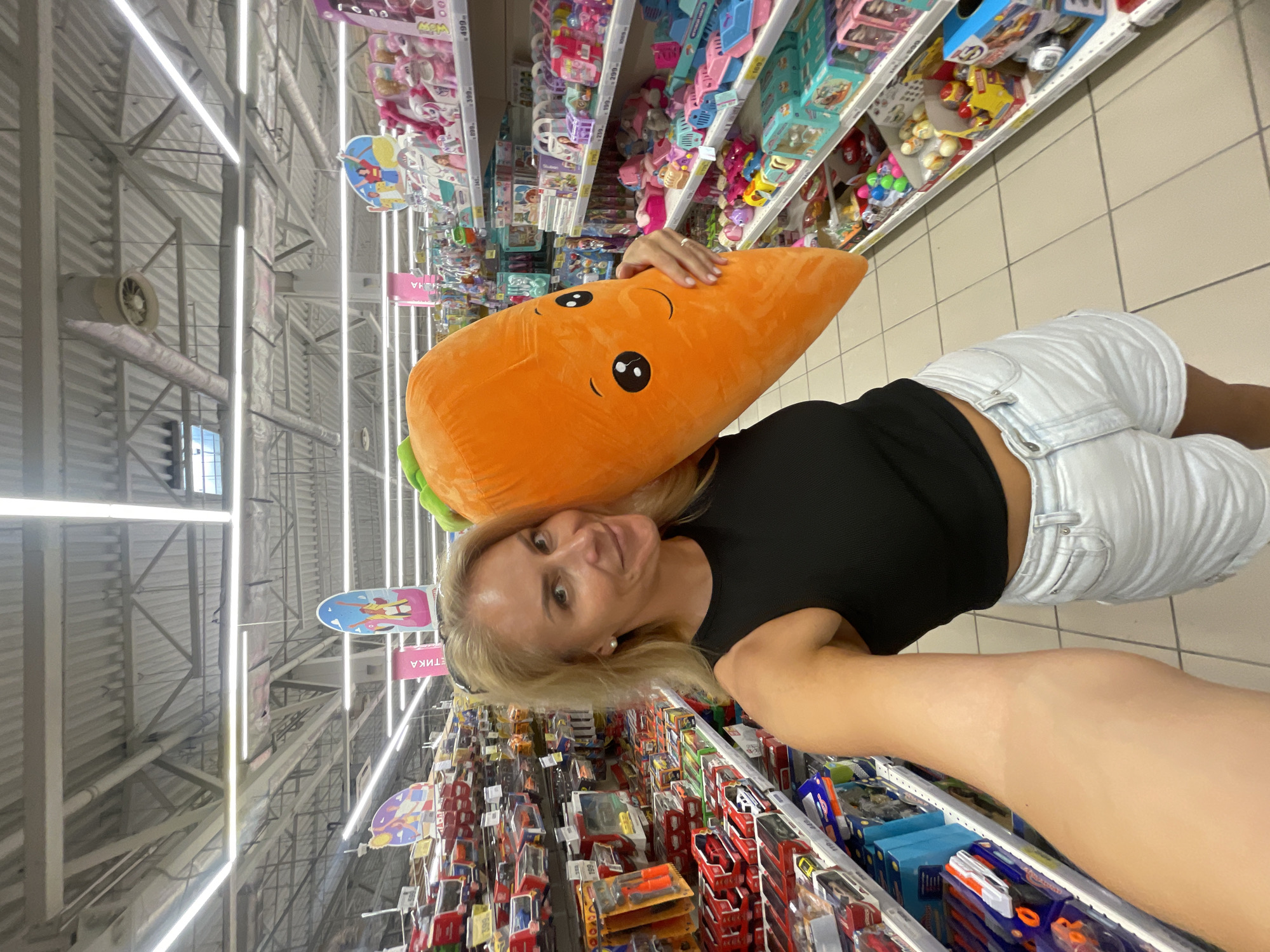 The image captures an adult female taking a selfie in a brightly lit supermarket aisle in Sochi, Russia. The woman, appearing to be in her 30s or 40s with blonde hair, is smiling and looking towards the camera. She is wearing a black sleeveless top and light-colored shorts. A pearl earring is visible in her right ear, and she wears a ring on her left ring finger. She holds a large, orange, cartoonish carrot-shaped plush toy with a green top and a smiling black-and-white face, positioned over her right shoulder.
The setting is a wide retail aisle within a store, with shelves extending into the background on both sides. The shelves on the left are stocked predominantly with packaged toy vehicles, including model cars in blister packs and boxes, and a blue and orange toy gun. Price tags displaying "199" are visible. The shelves on the right display a variety of children's toys, including dolls, small animal figures, and colorful plastic play sets. Price tags indicate values such as "899," "399," "499," "270," and "169." A prominent pink vertical banner with white Russian text, including "ЭТИКА" (ETIKA), hangs above the right aisle. Circular decorative signs with cartoon imagery are also suspended from the ceiling.
The ceiling above is high, featuring an exposed industrial structure with white corrugated metal roofing, metallic support trusses, and numerous long fluorescent light fixtures running the length of the aisle. Several circular ventilation ducts and diffusers are also visible. The floor is tiled with large, light-colored square tiles and dark grout lines. The presence of Russian text on the signage and price tags denominated in Rubles confirms the location within Russia, specifically Sochi as per the prompt.