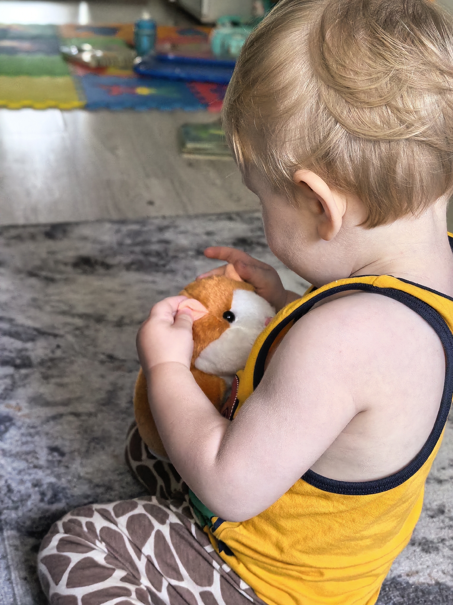 A toddler with light brown hair is sitting on a gray rug, holding a stuffed animal. The toddler is wearing a yellow tank top with black trim and brown and white patterned pants. The stuffed animal appears to be a fox or similar creature. In the background, a colorful play mat is visible on a wooden floor. The image is a close-up shot, focusing on the toddler and the toy. There are no discernible landmarks or indications of Sterlitamak, Russia.