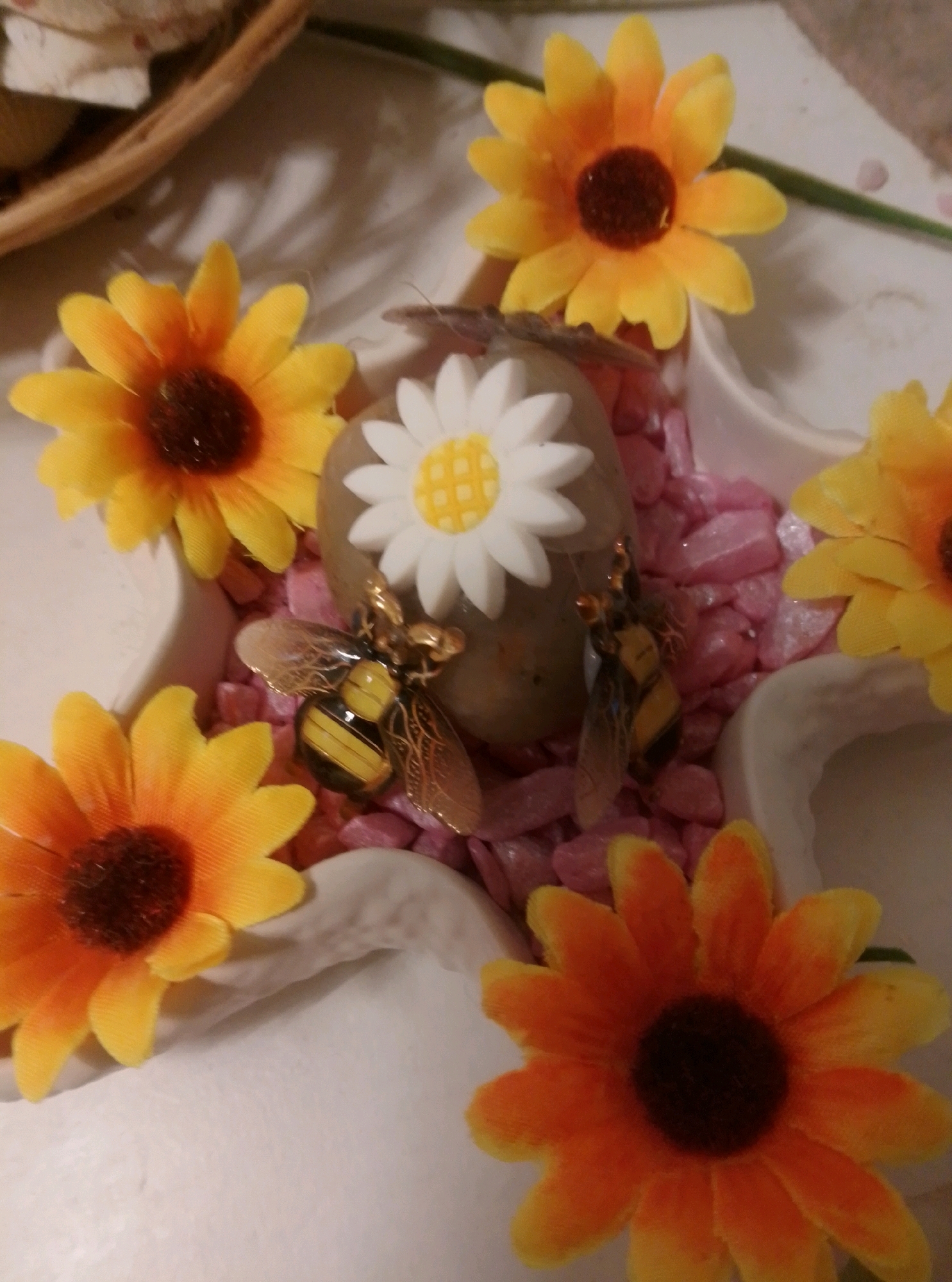 A close-up, overhead shot shows a decorative arrangement of artificial sunflowers, two bee figurines, and a single white daisy figurine nestled amongst pink gravel. The arrangement is presented in a white, petal-shaped bowl. The background is blurred, but appears to be an indoor setting with hints of a woven basket and a green stem in the upper right corner. There are no people visible in the image. The location in Wedel, Germany, is not identifiable from the visual content.