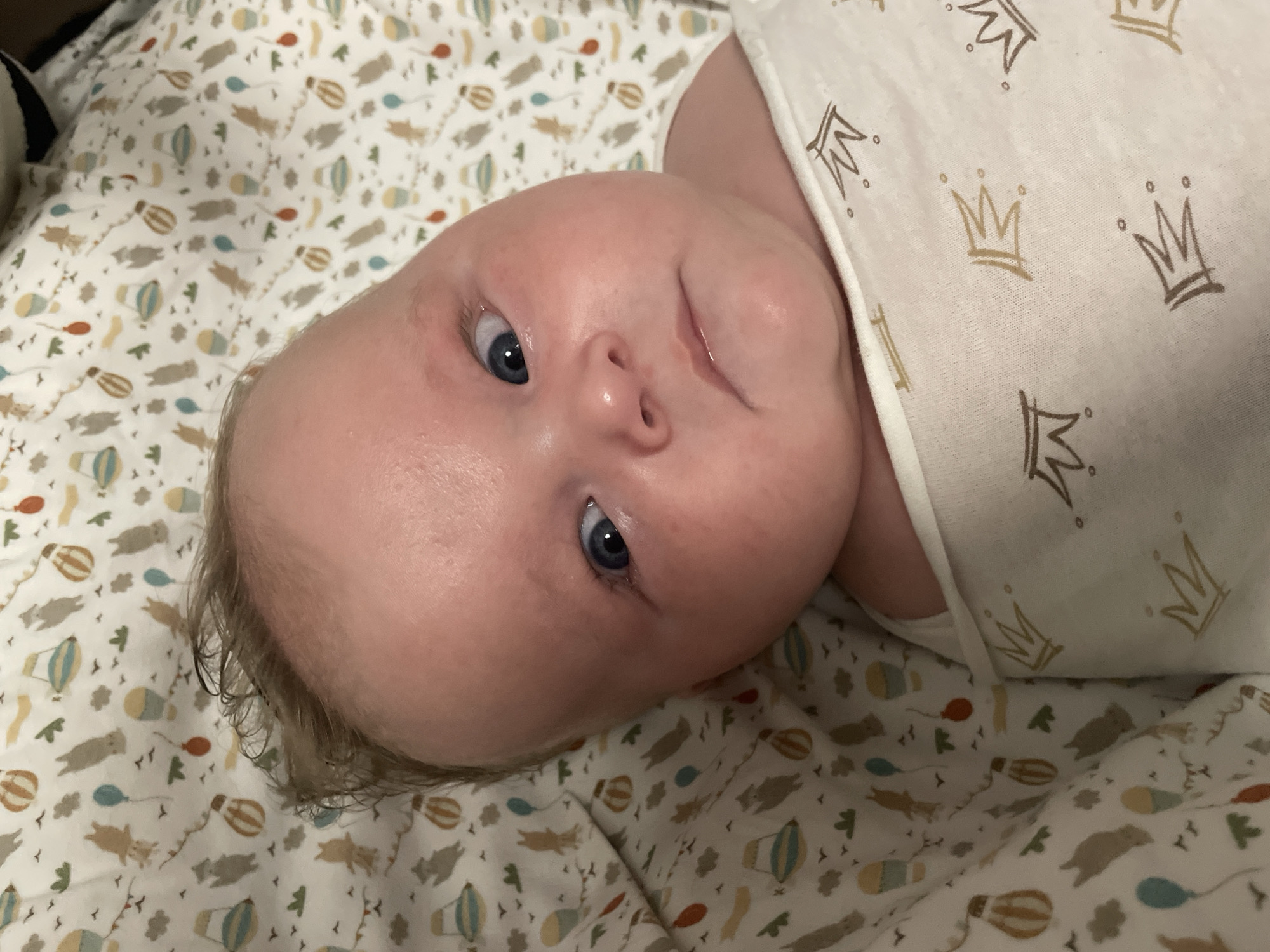 A close-up overhead shot shows a baby's face and upper body. The baby has fair skin, light brown hair, and blue eyes looking slightly upwards and to the left. The baby's left cheek appears slightly red. The baby is wrapped in a white swaddle blanket with a pattern of gold crowns and small dots. The background is a white fabric with a repeating pattern of cartoon animals, hot air balloons, and birds. There is no indication of the city of Stavropol, Russia in the image.