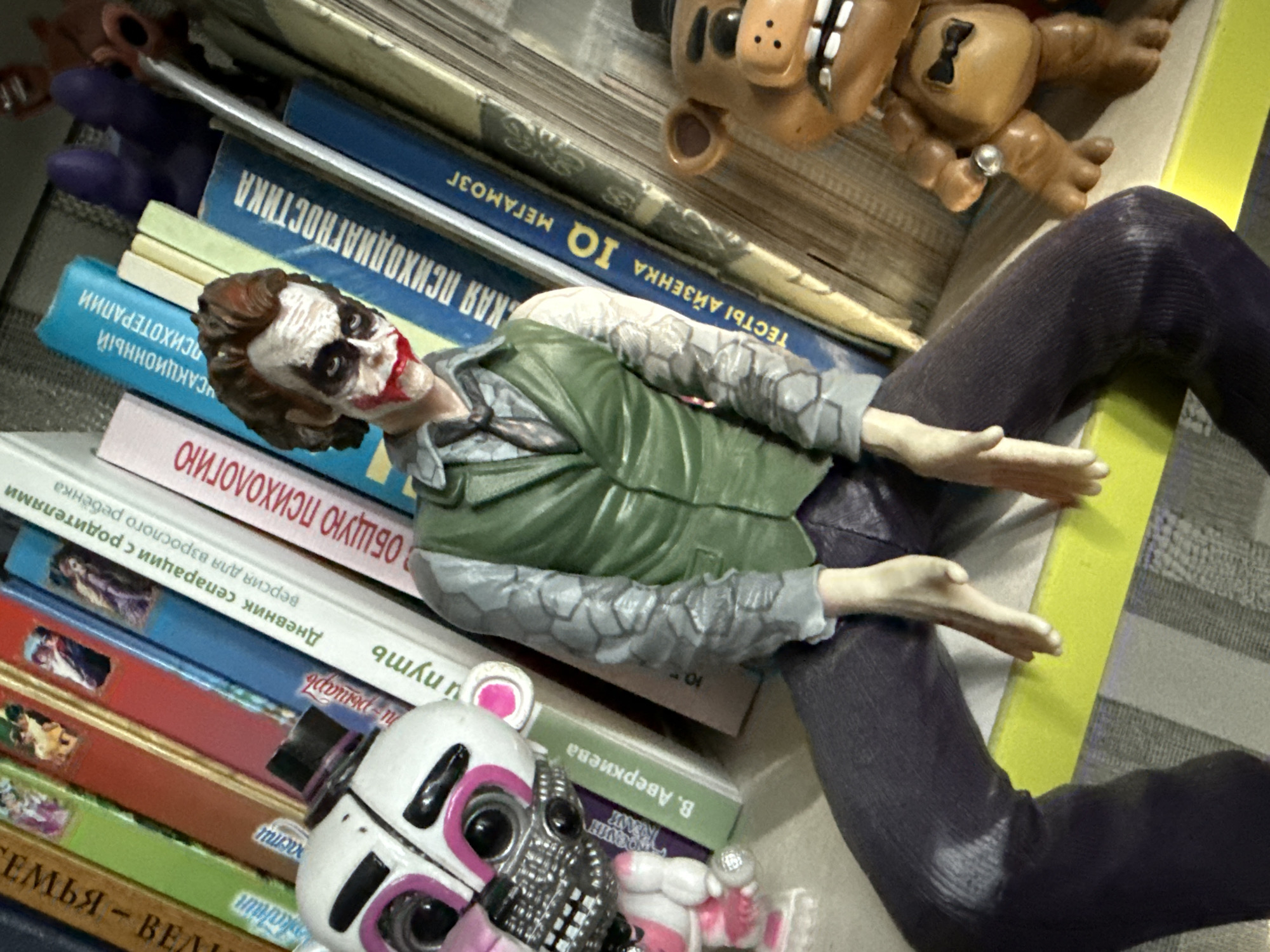 A close-up shot shows toy figures positioned in front of a shelf of books. On the left, a white and pink animatronic figure with black and pink accents is visible, partially obscuring the lower portion of a mannequin's torso, which is painted to resemble the Joker. The Joker mannequin is wearing a green vest over a grey shirt and tie, and dark pants. Its face is painted white with black around the eyes and red lipstick. To the right of the Joker, a brown bear-like toy figure with a black bowtie is present. The books on the shelf appear to be mostly non-fiction, with titles in Russian, including "Общая психология" (General Psychology), "Дневник сепарации с родителями" (Diary of Separation from Parents), and "Тесты Айзенка IQ Мегамозг" (Eysenck IQ Tests Megamind). The environment is a domestic setting, likely a room within a residential building. There is no indication of the specific location within the city of Simferopol, Ukraine, as the image lacks any identifying landmarks or contextual clues.