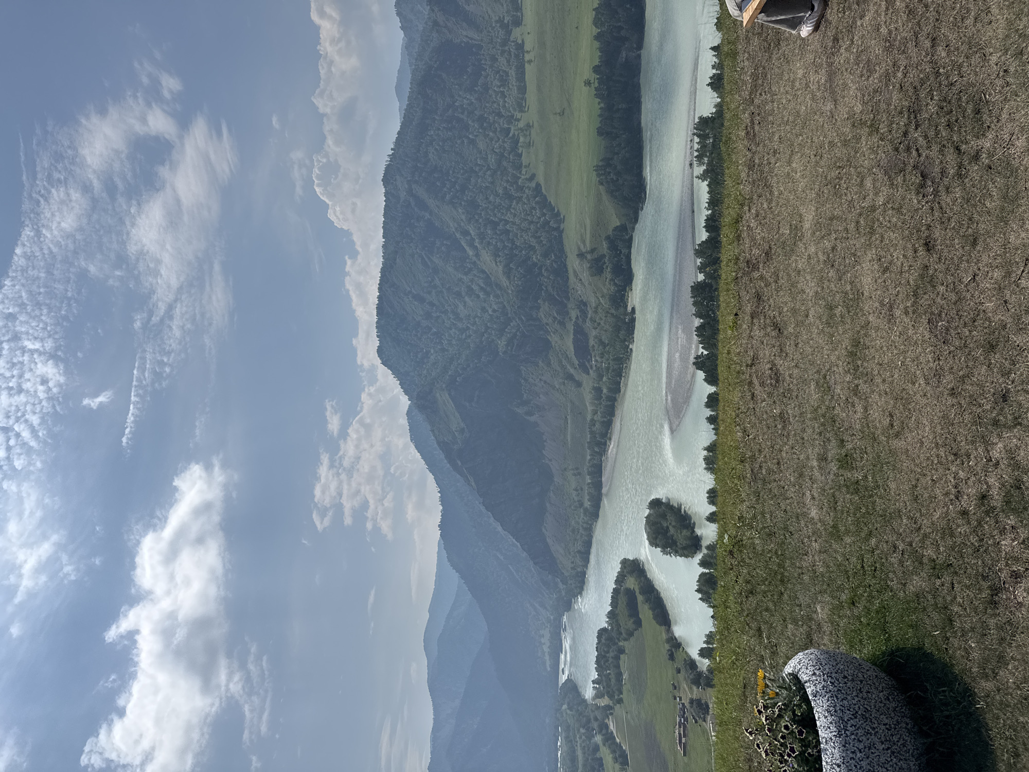 This image captures a sweeping panoramic view of the Katun River in the Chemal region of Russia. In the foreground, a large, textured stone planter filled with small flowers sits on a grassy knoll. The knoll slopes down towards the wide, turquoise Katun River, which meanders through a lush valley. On the far side of the river, steep, tree-covered mountains rise majestically, their peaks fading into a hazy blue in the distance. The sky is a bright blue, dotted with fluffy white clouds, casting dappled sunlight on the landscape. The scene conveys a sense of natural beauty and tranquility.