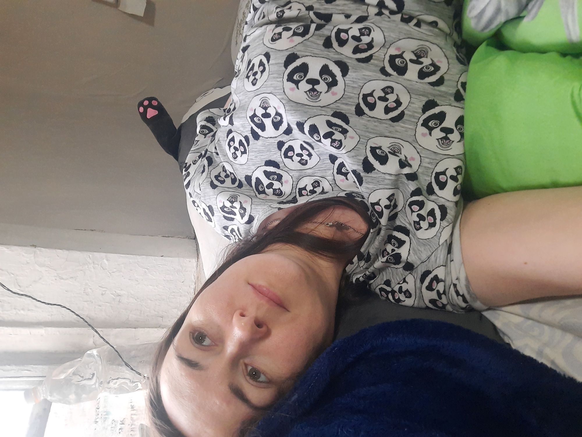 The image shows a woman lying down, wearing a gray pajama top with a panda bear pattern. She is resting her head on a dark blue blanket or pillow. Beside her is a bright green pillow. There is a black stuffed animal arm with pink paw pads visible in the background, appearing to be attached to a larger stuffed animal or pillow. The background shows what appears to be a window with a wire and a plastic bottle nearby. The location is possibly in Krasnogorskoye, Russia, but this cannot be definitively confirmed from the image alone. No specific actions or interactions are depicted.