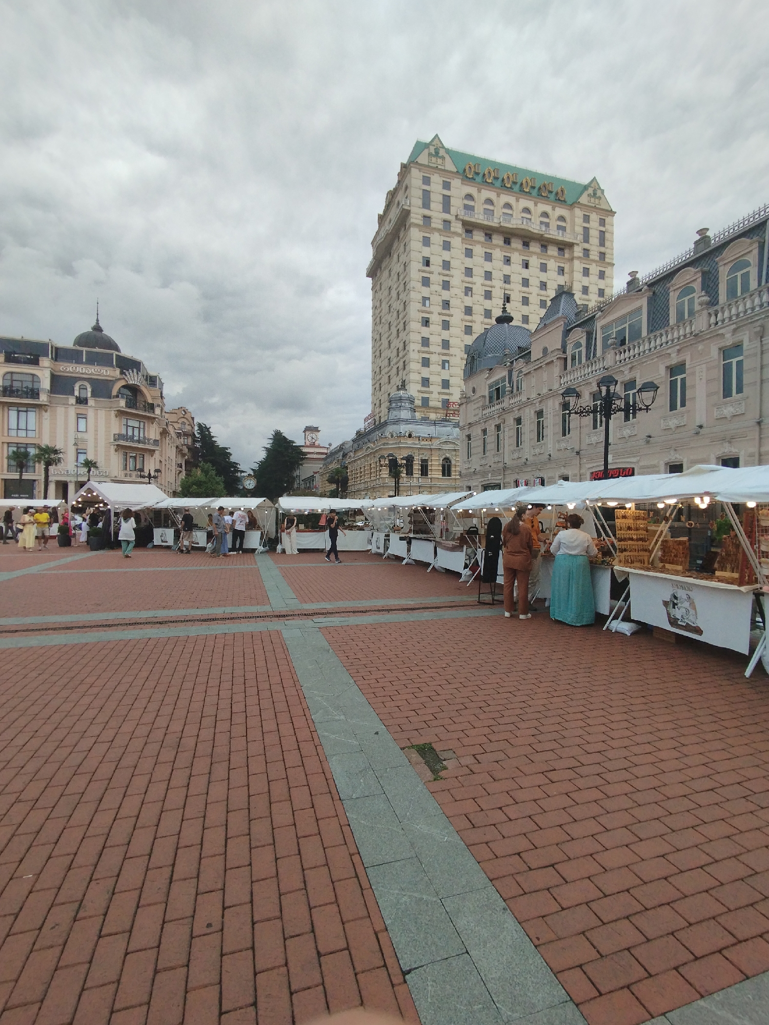 The image depicts a public square in Batumi, Georgia, featuring an open-air market under a cloudy sky. Several white tents are set up along the square, displaying goods for sale. People are browsing and interacting at the market stalls. In the background, a tall, light-colored building with a green roof stands prominently, along with other architectural structures characteristic of the city, including ornate facades and domes. The ground is paved with red bricks.