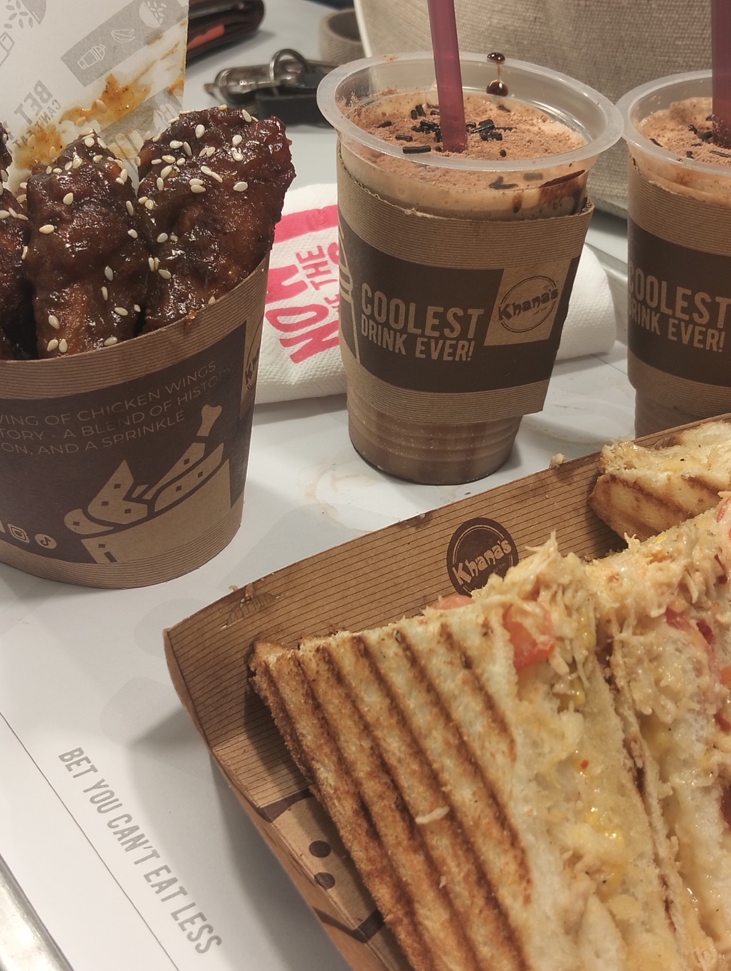 This close-up shot captures a casual meal set on a white surface, likely a table in a restaurant in Dhaka, Bangladesh. The foreground features two halves of a grilled sandwich, filled with a creamy mixture, corn, and diced red ingredients like tomatoes, resting in a brown cardboard tray bearing the "Khana's" logo. To the left, a brown paper cup holds several chicken wings coated in a dark, glossy sauce and generously sprinkled with white sesame seeds. This cup's text reads, "VING OF CHICKEN WINGS", "TORY - A BLEND OF HISTORY", and "ON. AND A SPRINKLE", along with social media icons. Below this, partially inverted text on the table says "BET YOU CAN'T EAT LESS".
In the background, two plastic cups contain cold beverages, likely chocolate milkshakes or iced coffees, topped with cocoa powder and chocolate sprinkles, each containing a red straw. These cups are encased in brown sleeves printed with "COOLEST DRINK EVER!" and the "Khana's" logo. A crumpled white napkin with the partial text "NOW" and "THE" is also visible. The scene depicts a prepared meal, possibly from a fast-food establishment, ready for consumption.