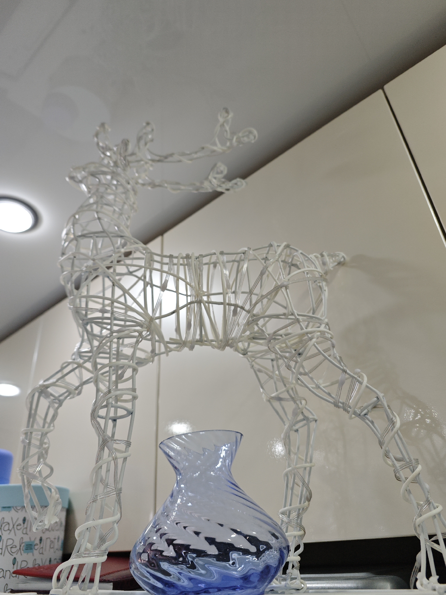 A low-angle shot captures a white, illuminated wire-frame reindeer decoration standing on a surface, likely indoors. The reindeer, intricately constructed from white wire wrapped with tubing, features subtle internal lighting that casts a soft glow from its body. Below the reindeer, to its right, sits a small, translucent blue glass vase with a swirling pattern. To the left of the reindeer, part of a light blue and white box or container is visible, displaying fragmented text that includes "axe", "Rele", and "nd", possibly hinting at words like "relax" or "release".
The background features light-colored wall panels or kitchen cabinets, with bright, circular recessed light fixtures visible on the ceiling. The objects appear to be placed on a dark countertop or shelf, which is seen from below. The overall setting suggests a domestic interior, possibly a kitchen or living area, decorated for a festive occasion. The artificial lighting from the ceiling and within the reindeer indicates an indoor scene where the time of day is not distinguishable from natural light. The image captures a static display of decorative items in a home located in Фоки, Russia.