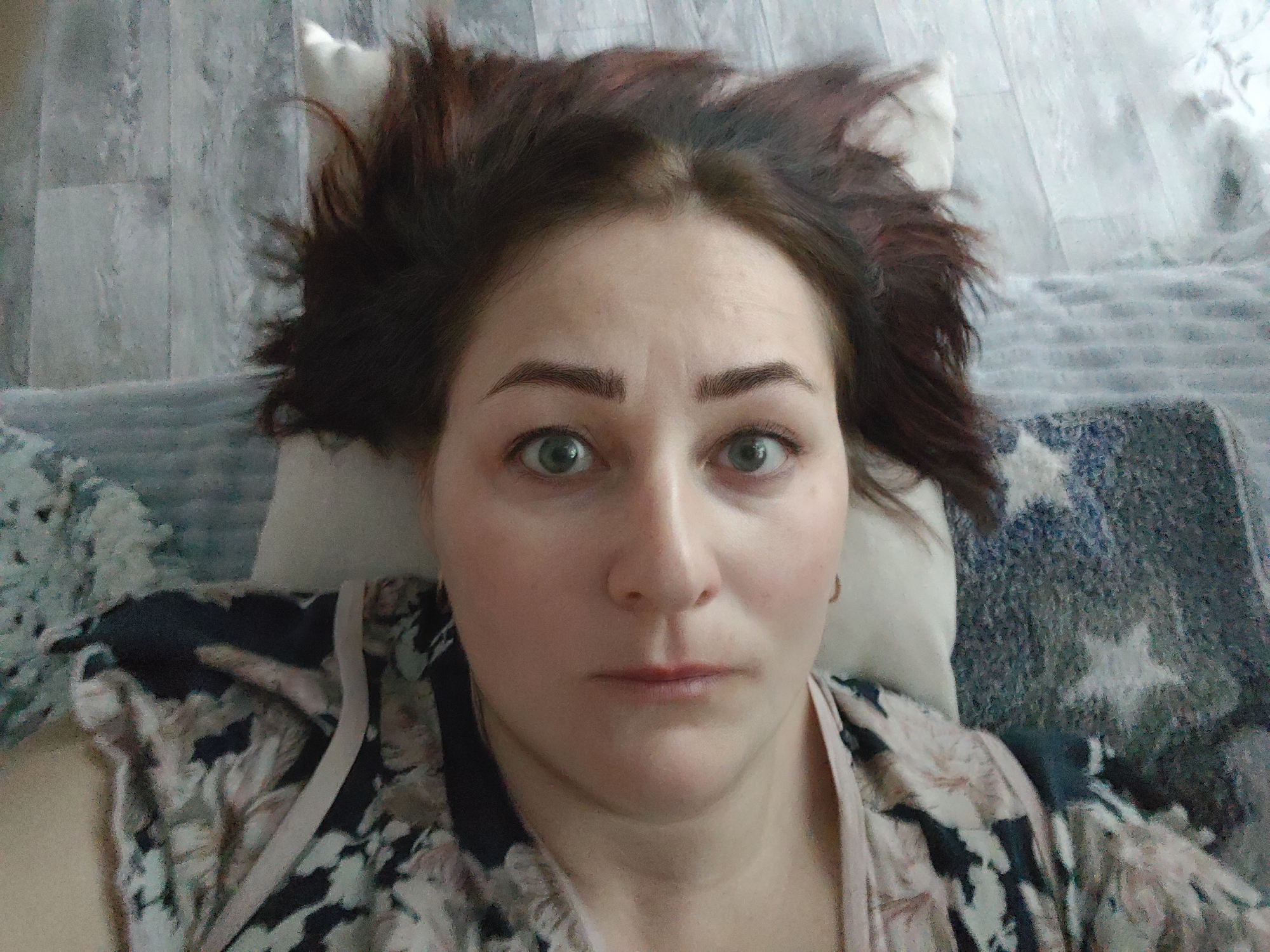 The image displays a close-up, overhead view of a single supine female individual in an indoor setting. The individual, appearing to be an adult, has dark brown hair, possibly with auburn highlights, fanned out and resting on a white pillow. Her face is pale with a neutral expression, wide-open light-colored eyes (possibly green or hazel) staring directly at the camera, and dark, shaped eyebrows. Her lips are closed with a faint reddish tint. She is wearing a garment with an abstract or floral pattern in black, beige, and light blue/white tones, with a light pink or beige strap visible near her right shoulder. To her right and below, a dark blue or grey patterned blanket with lighter star-like shapes is visible. The background above her head is a light-colored surface, possibly wood or tile with a textured finish. The scene is located in Markovskii, Russia.
