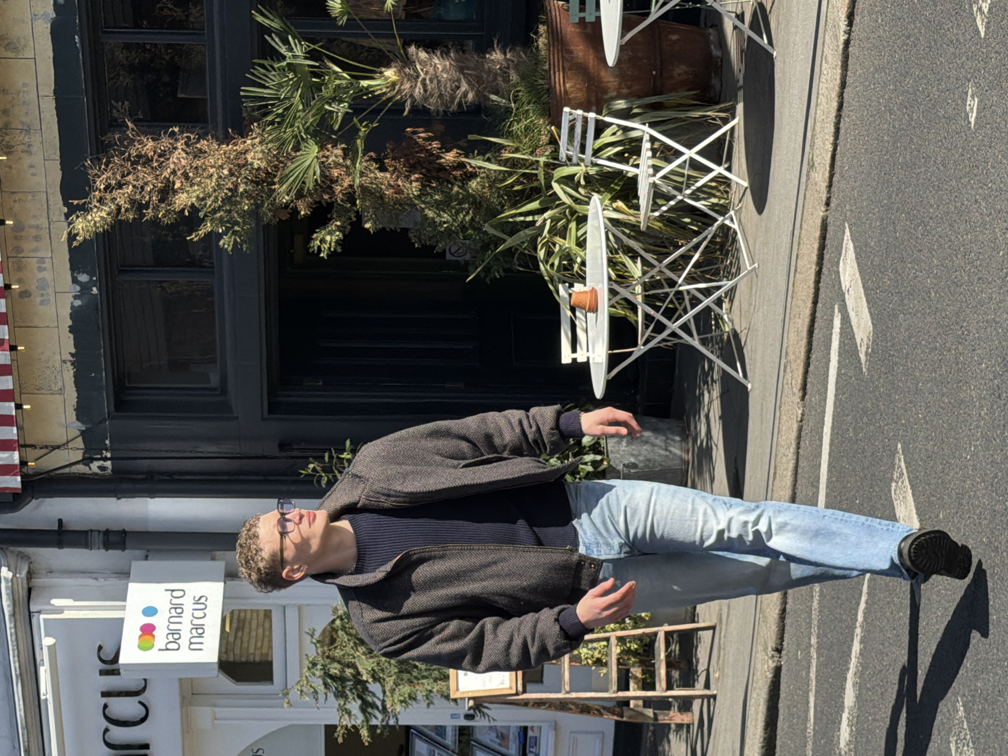 The image shows a man with short curly hair, wearing a dark jacket, a blue striped sweater, light blue jeans, and black boots, standing on a street. He is wearing glasses and looking slightly upward. Behind him is a building facade with black windows and a sign that reads "Barnard Marcus." Plants and outdoor tables are visible along the sidewalk in front of the building. The location is likely in London, United Kingdom, as Barnard Marcus is a real estate agency operating in that city. The street is paved and marked with white lines.