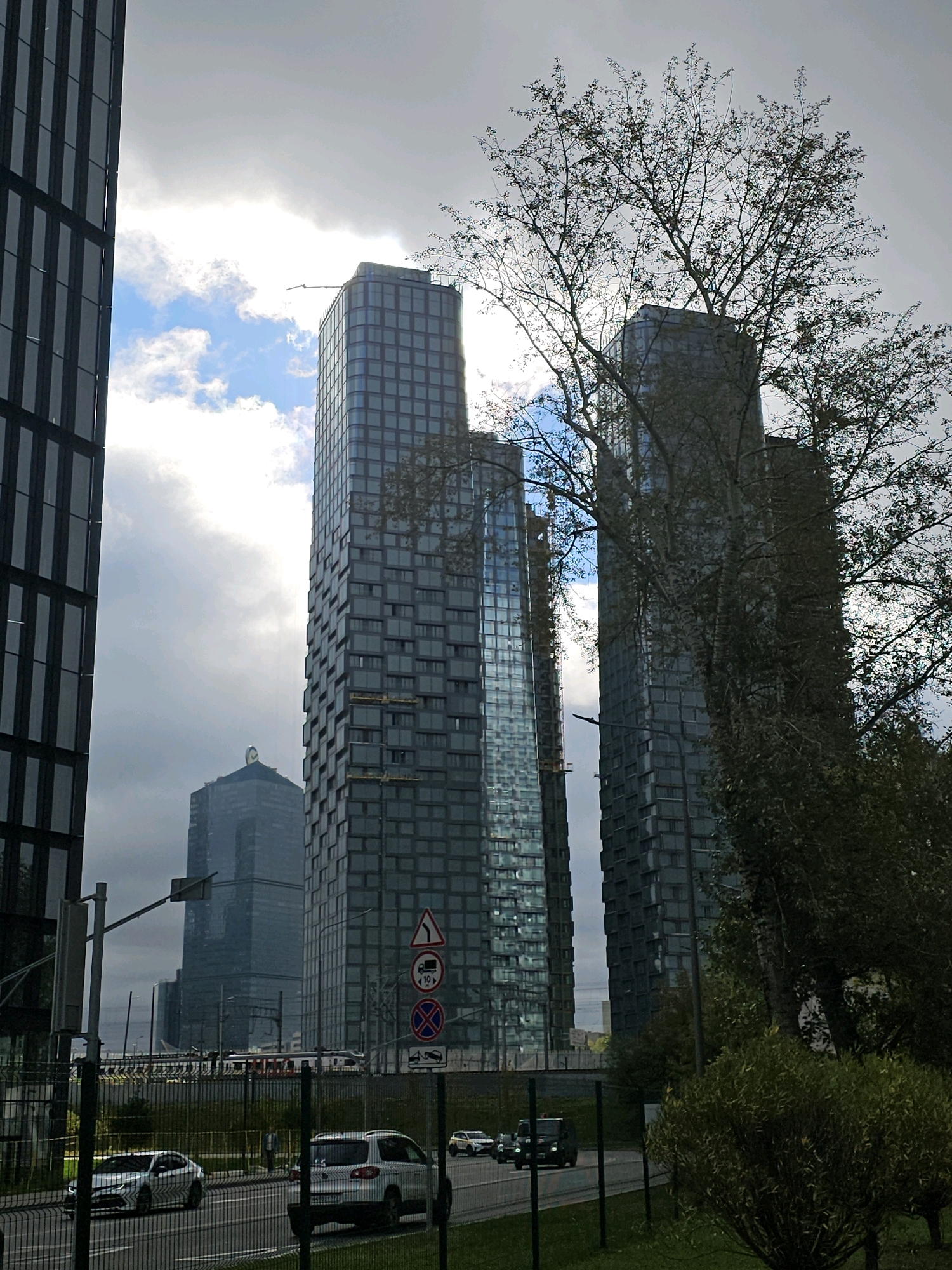 The image depicts a street view in Moscow, Russia, featuring several modern skyscrapers and vehicles. In the foreground, a black SUV is parked on the right side of a road, while a silver sedan is driving on the left. Behind them, a row of tall, glass-paneled buildings dominates the skyline. One of these buildings has a distinctive green-capped spire. The sky is partly cloudy with bright sunlight breaking through, casting reflections on the glass facades of the buildings. A tree with bare branches partially obstructs the view of the skyscrapers on the right side of the image. Traffic signs, including a triangular warning sign and a circular prohibition sign, are visible on the right side of the road. A metal fence runs along the edge of the road, separating it from a grassy area. The overall scene suggests an urban environment in Moscow, likely a business district due to the presence of numerous skyscrapers.