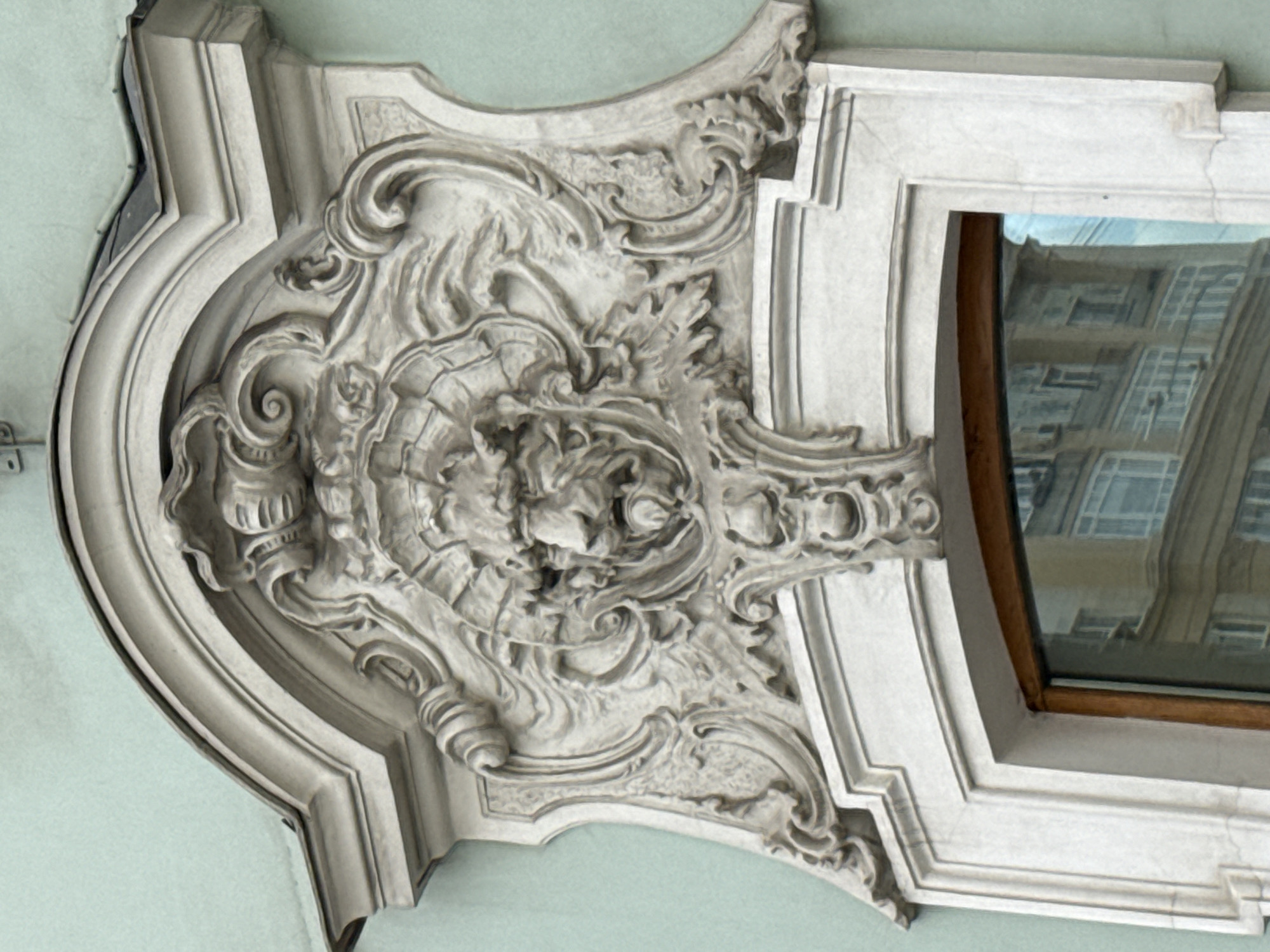The image shows a close-up view of an ornate architectural detail above a window on a building's exterior, likely in Moscow, Russia. The detail is a relief sculpture made of stone, featuring a central grotesque mask with a lion's face, surrounded by elaborate rococo-style ornamentation. The mask has a fierce expression, with bulging eyes, a prominent nose, and a wide, open mouth. The surrounding decorative elements include swirling acanthus leaves, scrolls, and volutes, all rendered in a highly detailed and dynamic manner. The sculpture is set within a curved pediment and framed by a stone architrave that forms the upper part of the window surround. The wall of the building is painted a pale green. Reflected in the window glass is a building with multiple stories and windows, suggesting an urban setting.