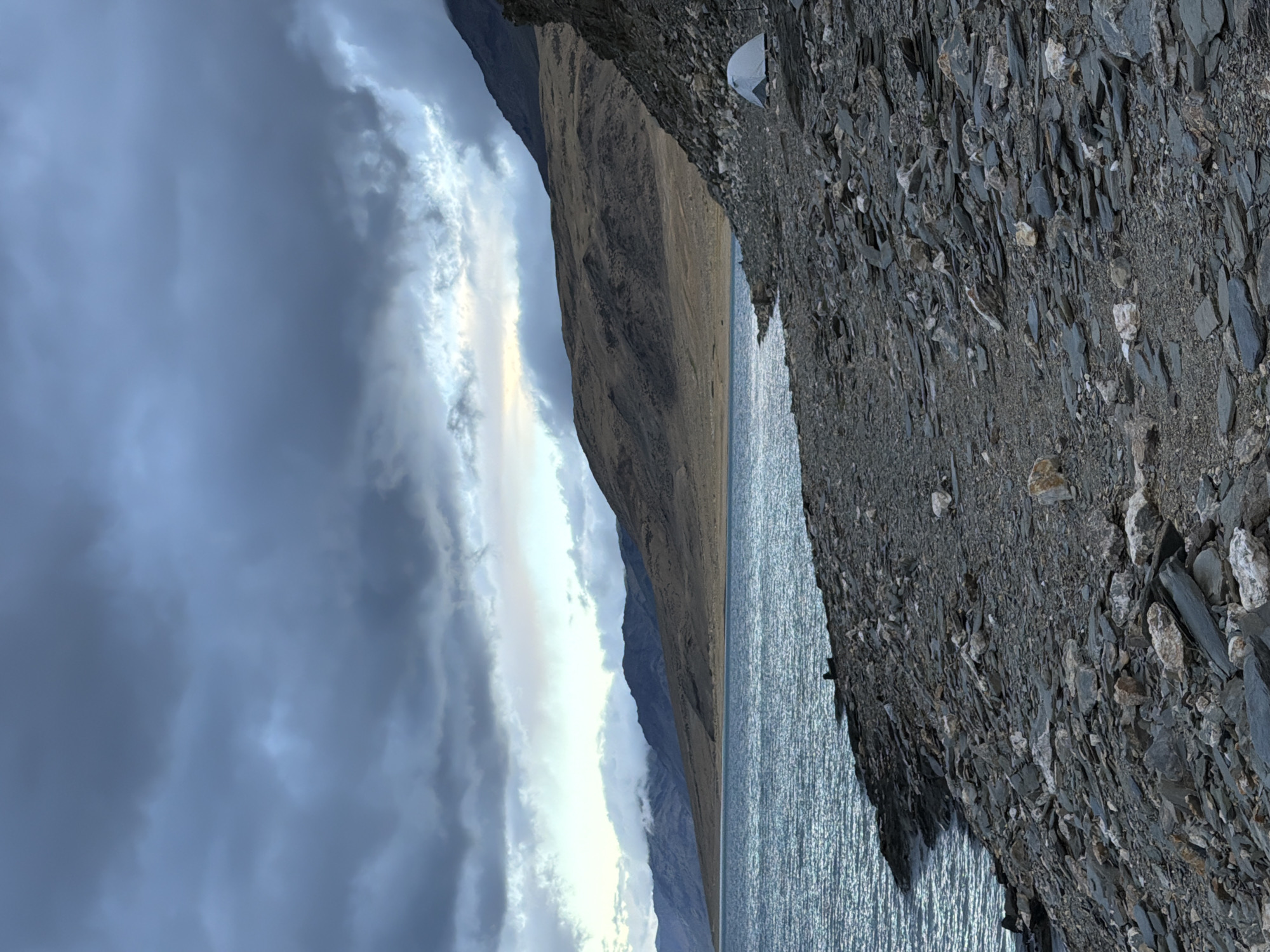 This is a vertical, medium-shot, outdoor photograph taken at dusk in Karzok, India. The scene depicts a rocky shoreline on the right side of the frame, with a body of water on the left. The water exhibits a turquoise hue and ripples, indicating a gentle breeze. The shoreline is densely covered with various sizes of grey and brown stones and rocks, interspersed with larger, lighter-colored rocks.
In the middle ground, a solitary white tent is pitched on the rocky shore, suggesting camping. Further back, barren, undulating hills with muted brown and tan vegetation dominate the landscape, leading to a range of darker, more rugged mountains in the distance. The sky is dramatic, filled with heavy, dark grey clouds that are partially illuminated by a soft, pale light breaking through in the upper center and right. This light casts a subdued glow on the distant mountains and the upper atmosphere. A faint, thin line of cloud seems to hug the peaks of the further mountains.
The overall mood is serene and slightly somber, characteristic of twilight in a remote, mountainous region. There are no visible people, animals, or vehicles in the image, apart from what appears to be a dark, silhouetted figure sitting on the rocks near the water's edge on the far left. The image captures a moment of solitude and natural beauty.