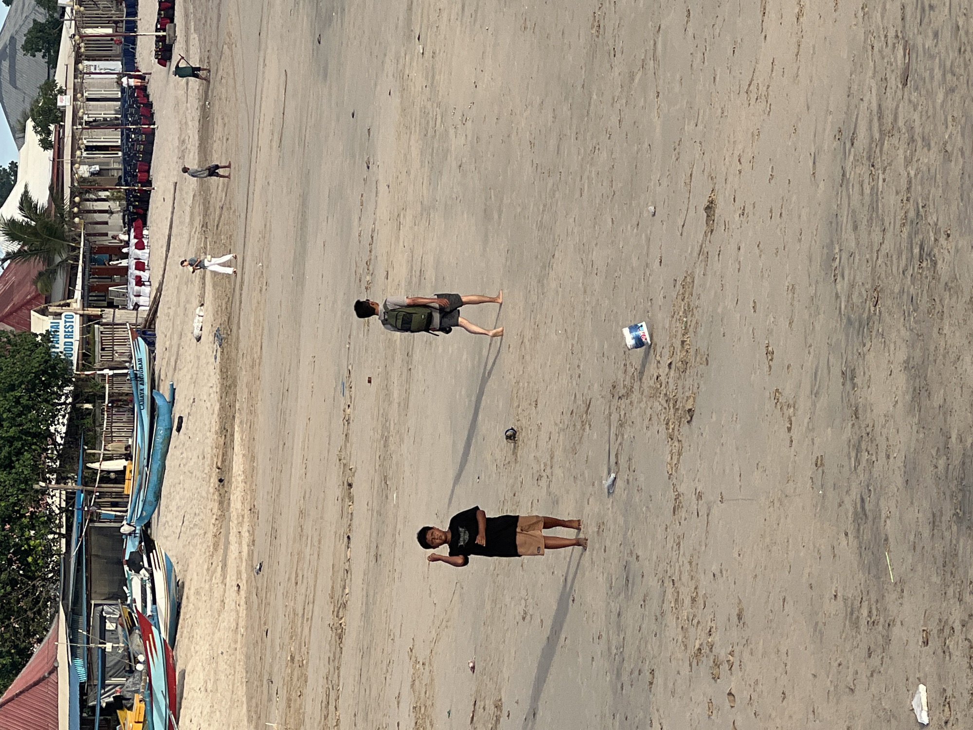 The image captures a scene on the beach in Kuta, Indonesia. The beach is sandy and fairly flat, with several people present. In the foreground, a man is lying down, while further up the beach, another man is walking with a backpack. In the background, there's a row of buildings and trees, which likely make up the resort area. Additionally, several fishing boats are docked near the trees. The scene is washed with sunlight, indicating a bright day.