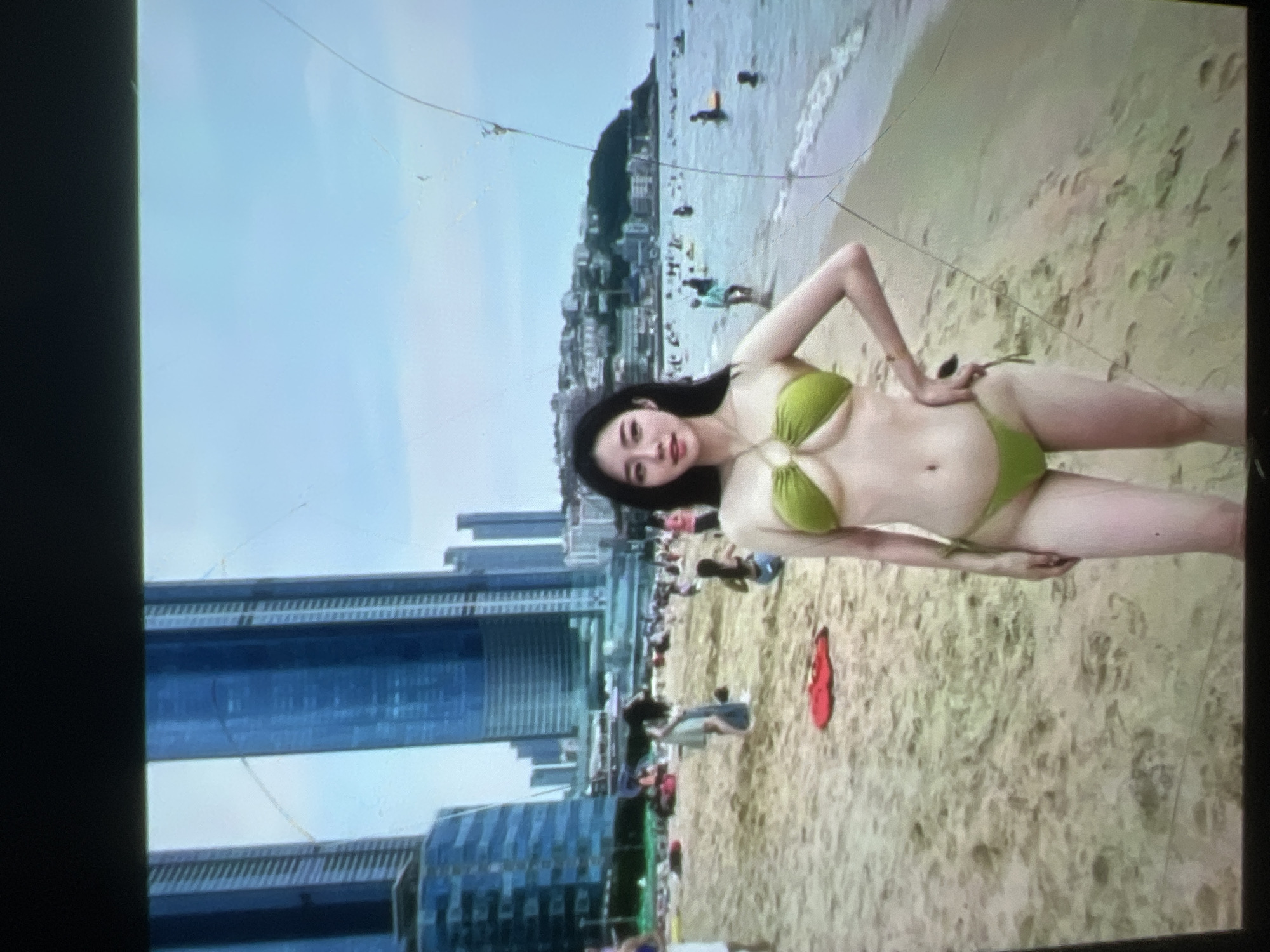 A young woman stands on a sandy beach wearing a lime green bikini. She has long black hair and is looking towards the camera with a neutral expression. Her hands are on her hips. In the background, the ocean is visible with several people in the water and along the shore. Beyond the beach, there is a cityscape with tall buildings, including skyscrapers. The sky is overcast. The image appears to be taken in an urban coastal area.