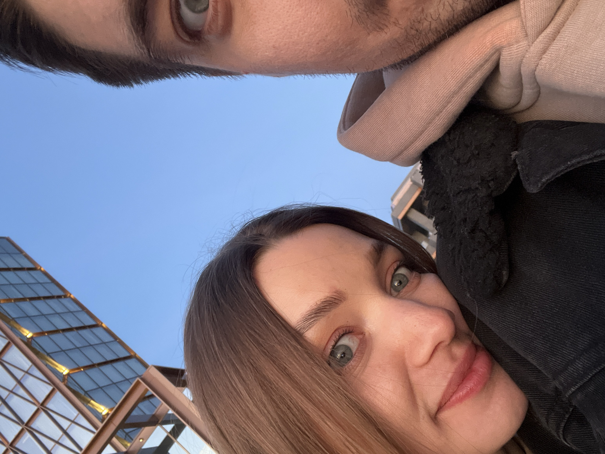 The image is an upward-angled close-up, likely a selfie, featuring two individuals against a clear blue sky and a modern building.
In the upper portion, a man's forehead, a green eye, and the bridge of his nose are visible, along with light stubble. He is wearing a light beige hooded sweatshirt, with the edge of the hood visible on the right.
The lower half of the image features a woman with long brown hair, prominent green eyes, and a gentle smile. She looks directly at the camera with an engaged expression. She is wearing a black denim jacket, revealing a black sherpa lining around the collar area.
The background is dominated by a bright, clear blue sky, suggesting daytime. To the left, part of a distinctive modern building is visible. It features large, geometric glass panels framed by light-colored metal structures, creating a grid-like pattern that slopes upwards. Some internal lighting or reflections are visible within the glass facade, indicating a contemporary architectural style. The overall impression is one of a casual, outdoor moment. No text is visible in the image.