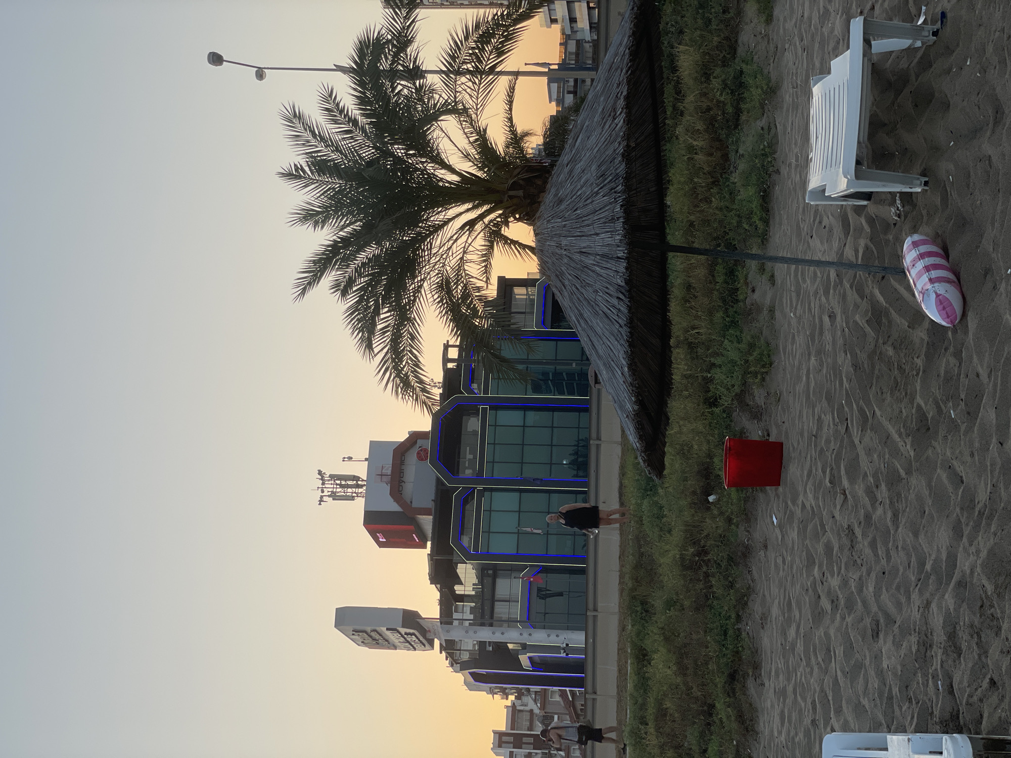 In Trikomo, Northern Cyprus, a sandy beach is visible in the foreground. A thatched umbrella shades a white plastic lounge chair and a pink and white striped inflatable on the sand. In the mid-ground, tall grass borders the beach. A red bucket sits amongst the grass, and a modern building with blue and white lights is visible behind it. A palm tree stands to the right of the building. Two people are walking on a path in front of the building. The sky is a pale orange, indicating either sunrise or sunset.