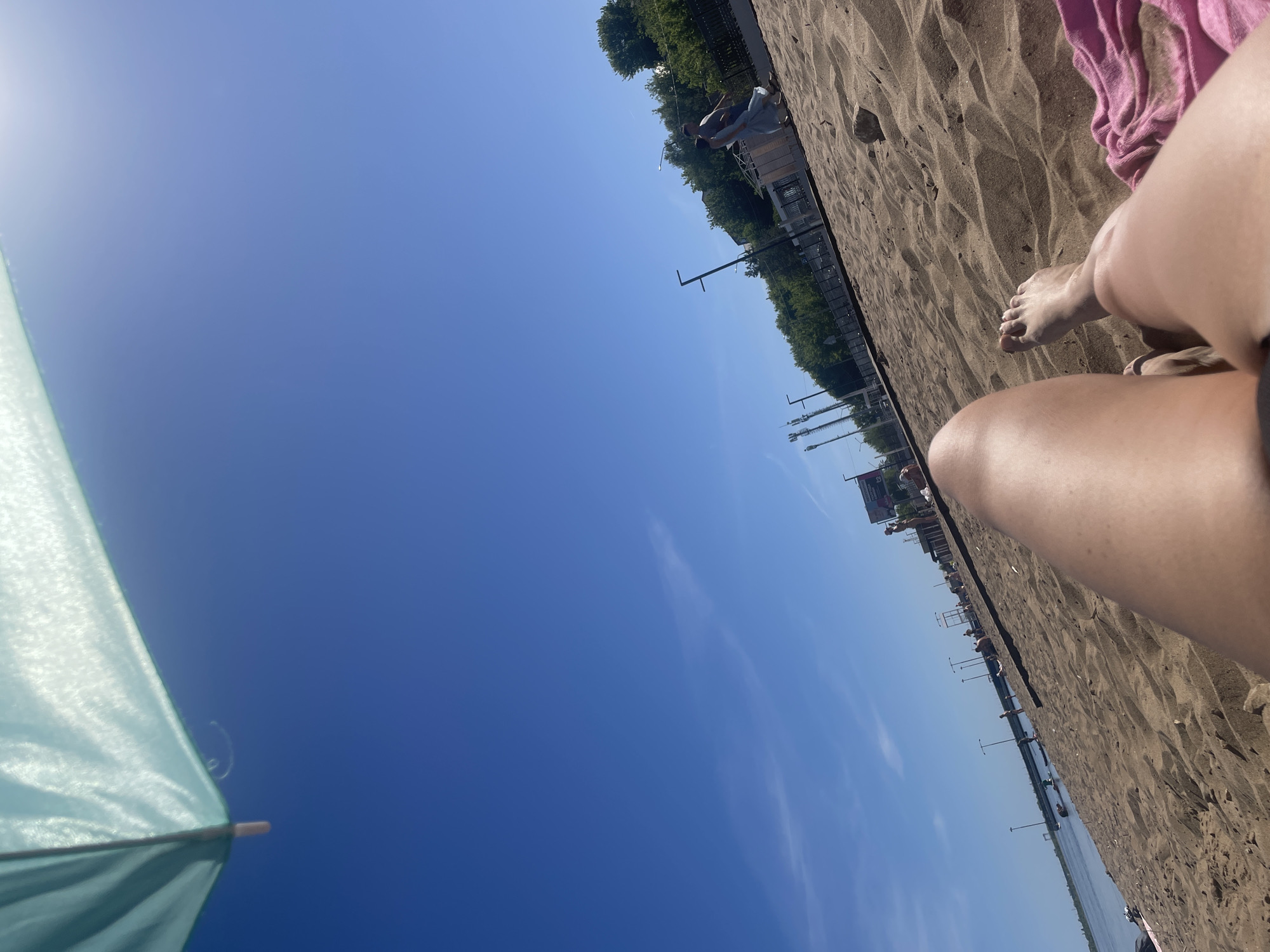 The image shows a beach scene in Saratov, Russia. A portion of a light blue beach umbrella is visible in the lower left corner against a clear blue sky. In the foreground, a person's legs are resting on the sandy beach. The sand appears to be coarse and textured. A pink towel lies nearby. In the distance, a row of streetlights lines the beach, and buildings and trees are visible. The water's edge is barely visible in the distance. The photo is taken from a low angle, looking upwards.