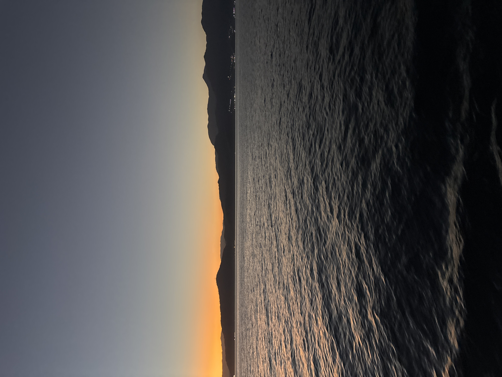 The image depicts a serene coastal landscape at dusk. The sky transitions from a deep blue at the top to a warm orange glow near the horizon, suggesting the last moments of sunset. Silhouetted against the fading light are a range of mountains, their peaks softly defined. Along the base of the mountains, scattered lights indicate a town or settlement nestled by the coast.
The foreground is dominated by a body of water, likely the sea, with gentle ripples reflecting the sky's colors. The water appears dark and calm, with the light catching the crests of small waves. The overall impression is one of tranquility and natural beauty as day turns into night.
The location is the city of Angra dos Reis, Brazil, known for its stunning bay dotted with islands and its mountainous coastline.