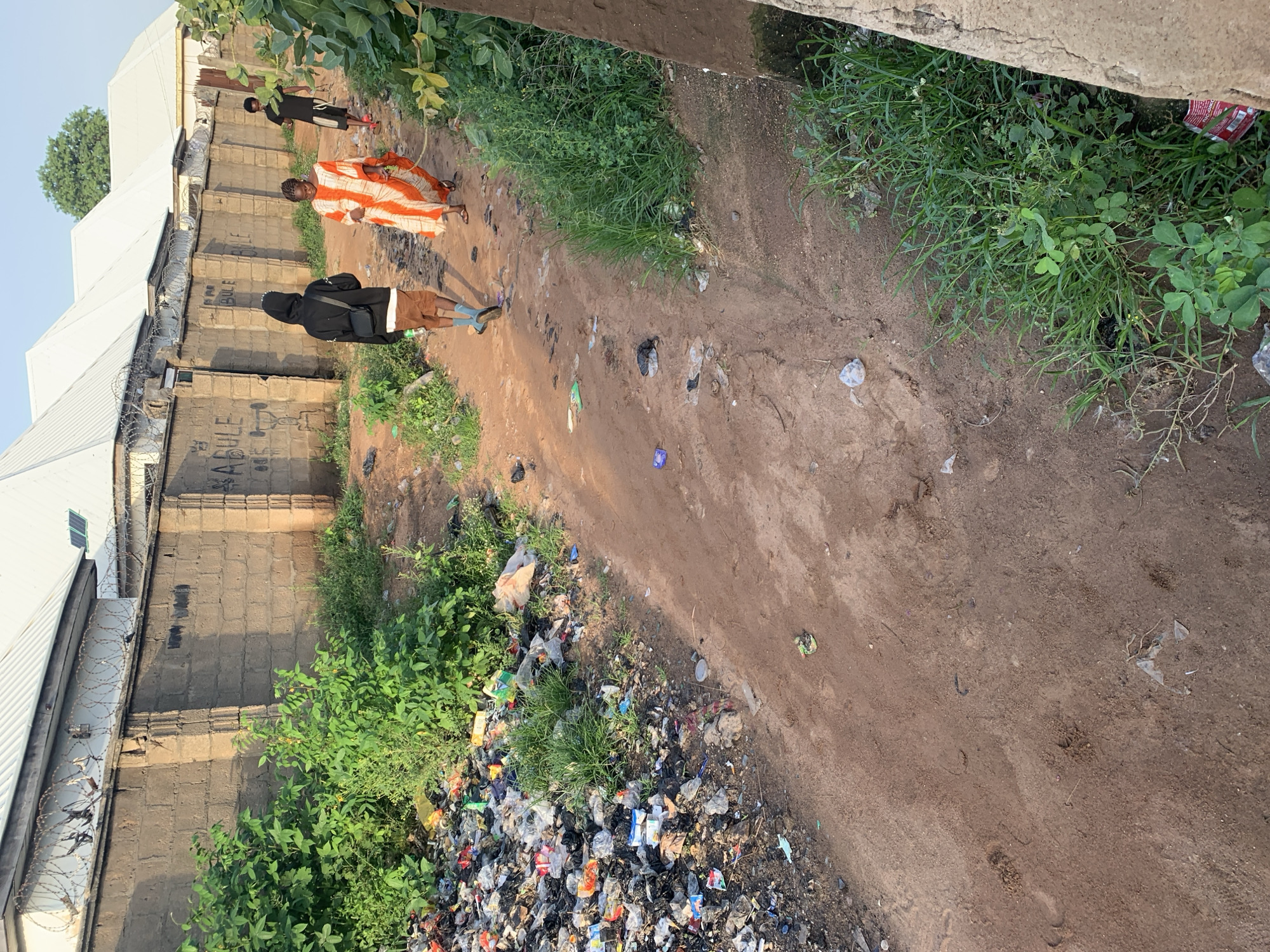 The image depicts a rural road in Jalingo, Nigeria, with a large pile of garbage on the left side of the frame. The garbage consists of various plastic bags, bottles, and other debris, with some green vegetation growing through it. The road itself is unpaved, made of red earth, and appears to be somewhat uneven. In the background, a stone wall with barbed wire runs along the property line of several buildings with white, corrugated iron roofs. Three individuals are walking away from the viewer down the road. The person in the foreground on the left wears a black hooded jacket and brown shorts, with blue socks and dark shoes. To their right, a person in the middle wears a flowing orange and white striped garment. Further down the road, a third individual, dressed in a black t-shirt and dark shorts, walks ahead. The environment is characterized by a clear sky and the presence of natural vegetation alongside the road and the garbage heap.
