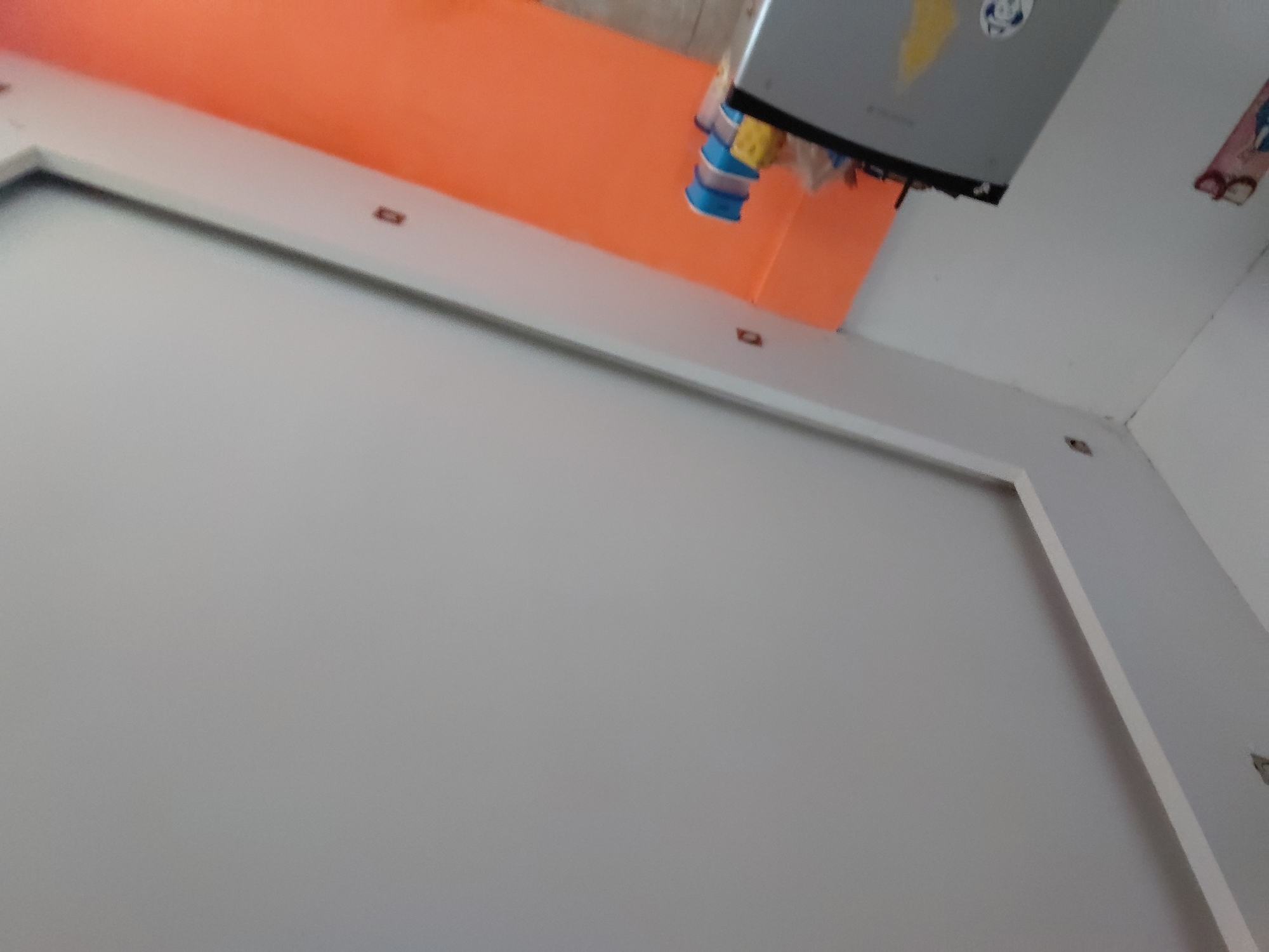 The image captures an upward view of an indoor setting, likely a room in Santa Ana, Venezuela, focusing on the ceiling and upper wall areas. No people are present in the scene.
A prominent, large, rectangular, gray electronic device, possibly an air conditioner or a projector, is mounted high on the wall. Several colorful items, including blue, yellow, and white fabric or plastic pieces, are seen hanging from its underside. A vibrant orange-colored wall or partition occupies a significant portion of the upper area beneath the mounted device. The ceiling and adjacent walls are painted a light gray or white.
A white, rectangular molding or frame runs along the junction of the ceiling and walls, featuring small, dark, rectangular inserts that could be fasteners or small embedded lights. A corner of the room with a structural joint is visible on the right.
No specific activity or event is taking place; the scene is static. The image is somewhat blurry and captured from a low, upward angle. Notable details include a yellow triangular sticker and an illegible circular logo on the visible surface of the mounted device. The overall impression is that of a casually decorated indoor space. No legible text is visible other than the indiscernible logo.