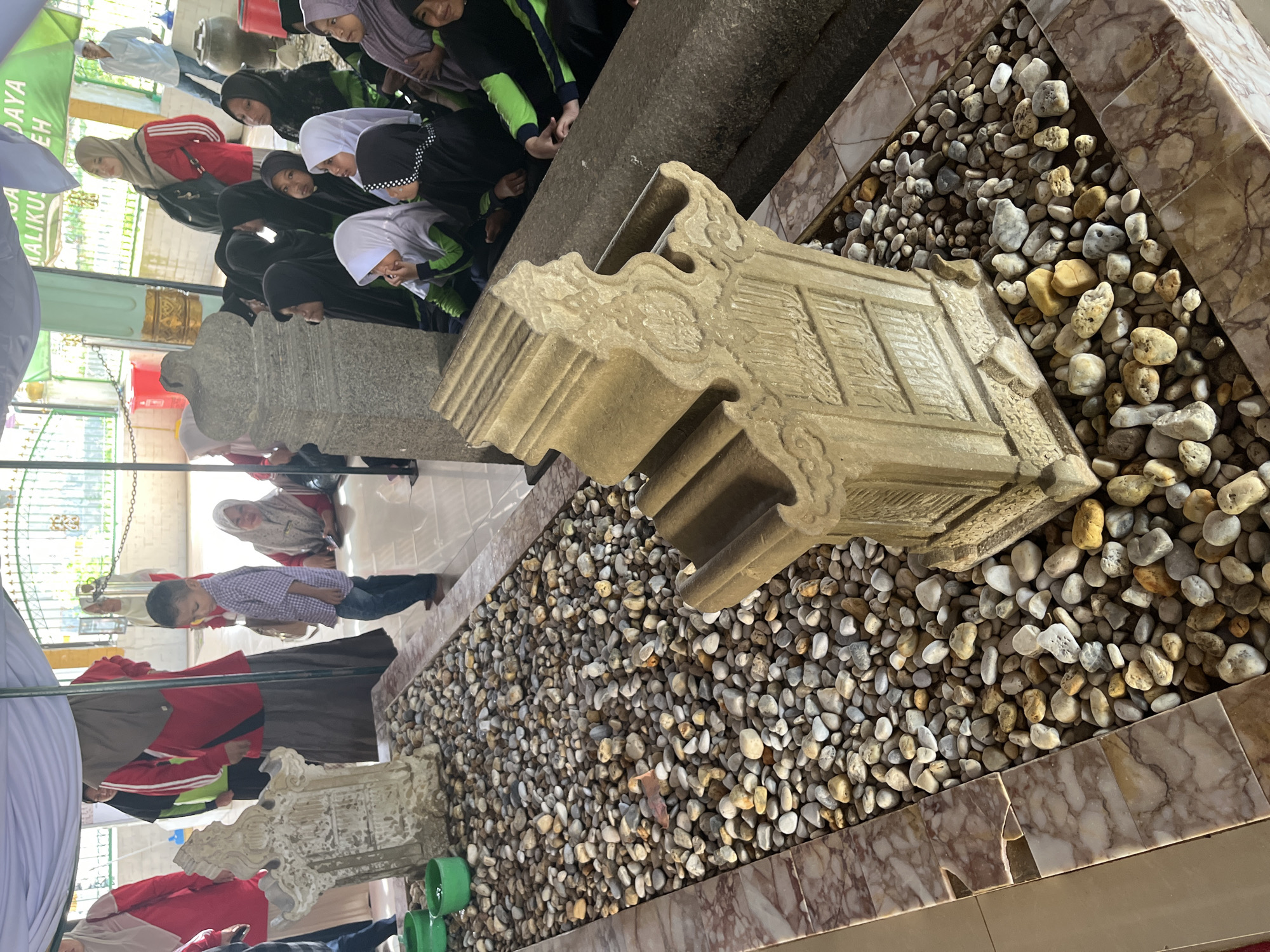The image depicts a scene at the Sunan Ampel Mosque complex in Beuringen, Indonesia. In the foreground, a sandstone gravestone, intricately carved with Arabic calligraphy, rests on a bed of small, multi-colored pebbles. To the left of the gravestone are three green bowls, also filled with pebbles. Behind the gravestone, a long, dark, stone sarcophagus is visible. A group of people, mostly women and children wearing hijabs, are gathered in the background, some sitting and observing the gravestones. A young boy in a plaid shirt stands between the gravestone and the group of onlookers. In the far background, a man in a white shirt and jeans stands near a large clay pot. The overall environment appears to be an outdoor, covered area, possibly a cemetery or a sacred site, with patterned marble tiling on the floor and a white canopy overhead. The lighting suggests it is daytime, with some areas in shadow.