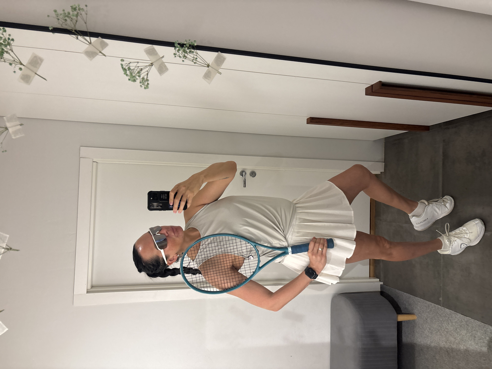 A woman is standing in a room, possibly a dressing room or a hallway, and taking a mirror selfie. She is wearing a light-colored tennis outfit consisting of a sleeveless top and a pleated skirt. She has on white tennis shoes with socks and a pair of white-rimmed sporty sunglasses. She is holding a teal tennis racket with a blue grip in her right hand. Her left arm is bent at the elbow, and her left hand is on her hip. A smartphone is in her right hand, used to take the photo. Her hair is dark and pulled back into a braid. The background shows a white door and a large mirror with some small green sprigs taped to it. To the left, a gray ottoman-like bench with wooden legs is visible. The floor appears to be dark gray tiling. The image was taken indoors. There is no visible indication that this location is in the city of Chaikovsky, Russia.