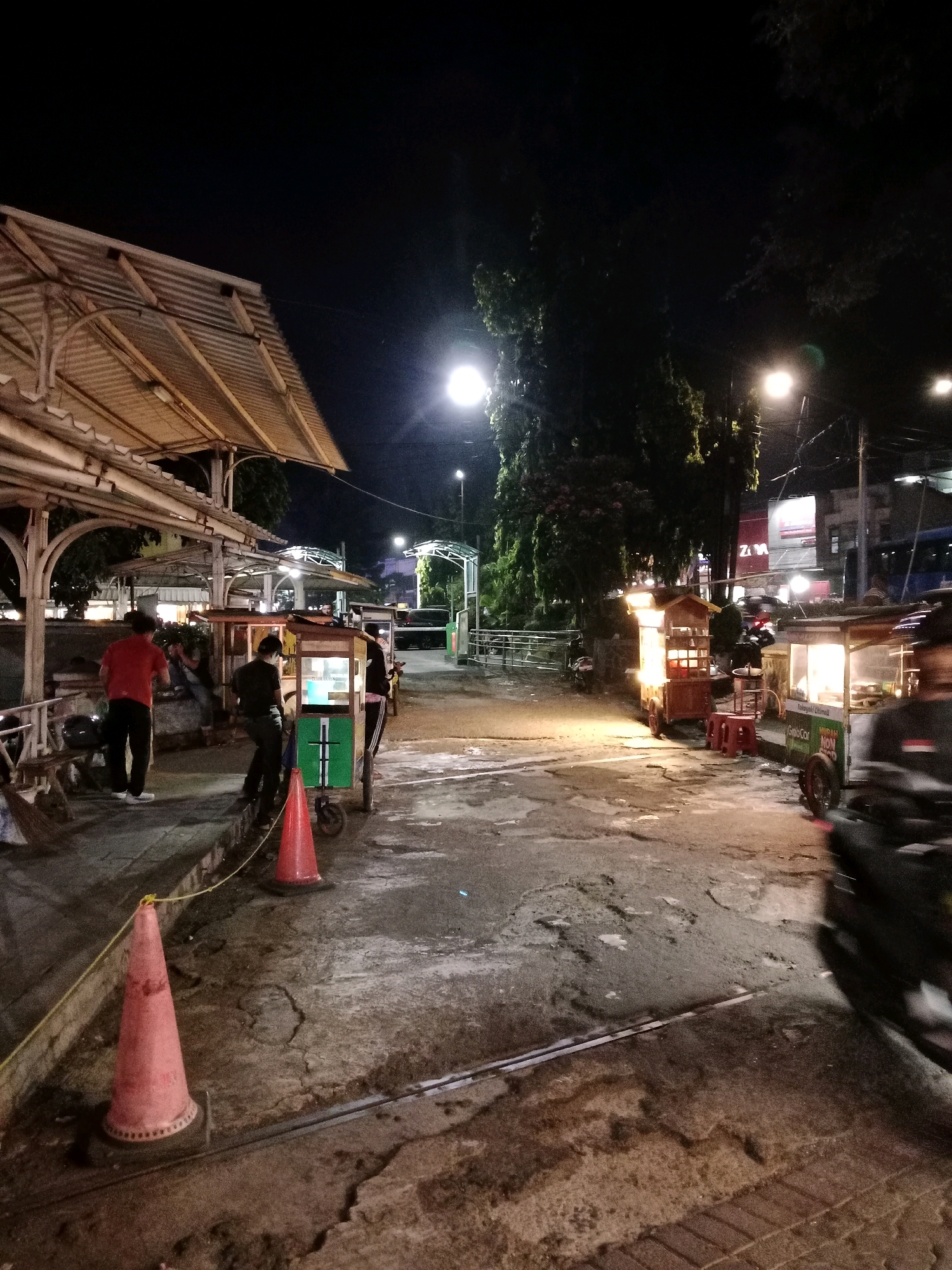 The image depicts a street scene at night, likely in Cianjur, Indonesia, with several food carts and people present. A covered structure with a corrugated metal roof is visible on the left, possibly a marketplace or seating area. Several individuals are standing near or operating food carts, which are illuminated and appear to be selling food. One person in a red shirt and black pants is standing near a cart, facing away from the camera. Another person in black is attending to a green and white cart. In the foreground, an orange traffic cone is placed on the ground. The street surface is asphalt with visible cracks and some water. Further down the street, more illuminated food carts are visible, along with trees and streetlights. The scene suggests a bustling night market or a street food vendor area. A blurry, fast-moving object, likely a motorcycle, is captured in motion on the right side of the frame.