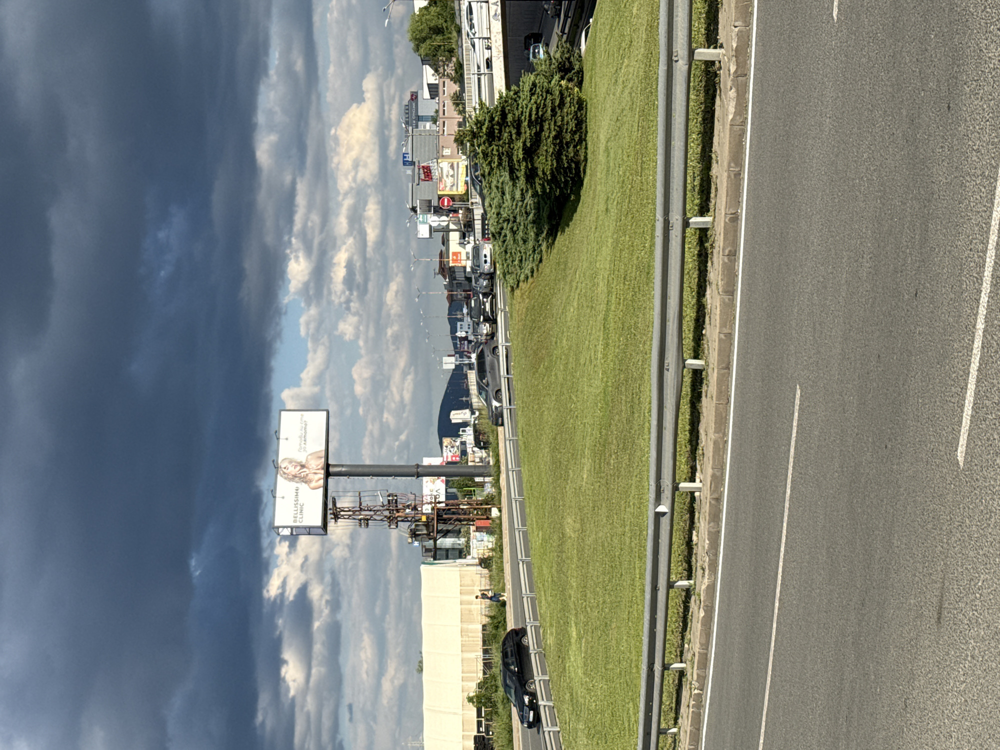 The image depicts a highway scene with a view towards the city of Sofia, Bulgaria. The sky is partially covered with dark storm clouds on the left side of the frame. A billboard advertising "BELLISSIMI CLINIC" is visible on the left side, in front of the sky. The right side of the frame is dominated by a two-lane highway with a metal guardrail. A grassy median strip runs between the highway lanes. A cluster of green shrubbery is observed growing in the median. In the distance, city buildings and signs are visible, along with heavy traffic. The road is surrounded by buildings, streetlights, and other signage. A car is parked on the shoulder of the road near the bottom left corner of the image. A person can be seen walking near the highway.