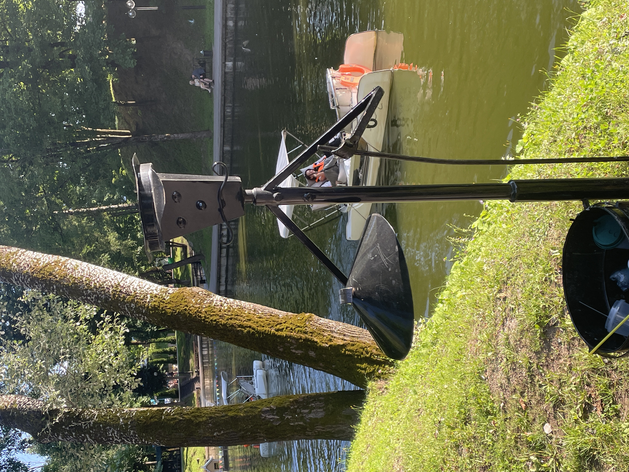 In Radkevshchina, Russia, a sunny day unfolds by a serene body of water. Two large trees with moss-covered trunks stand prominently in the foreground, their leaves casting dappled shadows on the lush green grass. Beyond them, the water reflects the clear blue sky and surrounding trees. A black bucket, containing what appears to be a yellow pole and some plastic containers, sits near the edge of the grass.
In the middle ground, a white paddle boat with a canopy is navigating the water. A person wearing an orange life vest is seated in the boat, steering it. Two orange buoys are attached to the back of the boat. Further in the distance, another white paddle boat is visible. On the far bank of the water, a paved walkway runs alongside a gentle grassy slope dotted with trees. Two individuals are seated on a bench on this walkway, enjoying the peaceful atmosphere. The overall scene depicts a tranquil recreational area in Radkevshchina on a pleasant day.