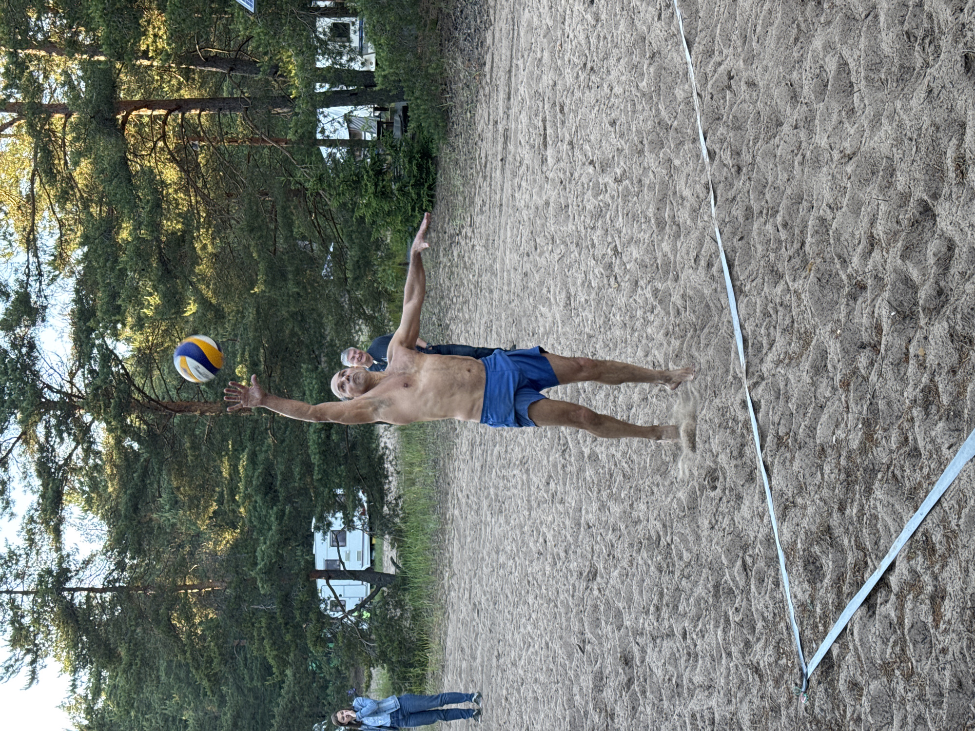 A shirtless man in blue shorts is playing volleyball on a sandy court. He is in mid-air, jumping to hit the ball which is above him. The court is marked with a blue line tape. In the background, there are trees, two white campers, and two other people. The scene appears to be in Zelenaya Roshcha, Russia, a resort town known for its pine forests and beaches.