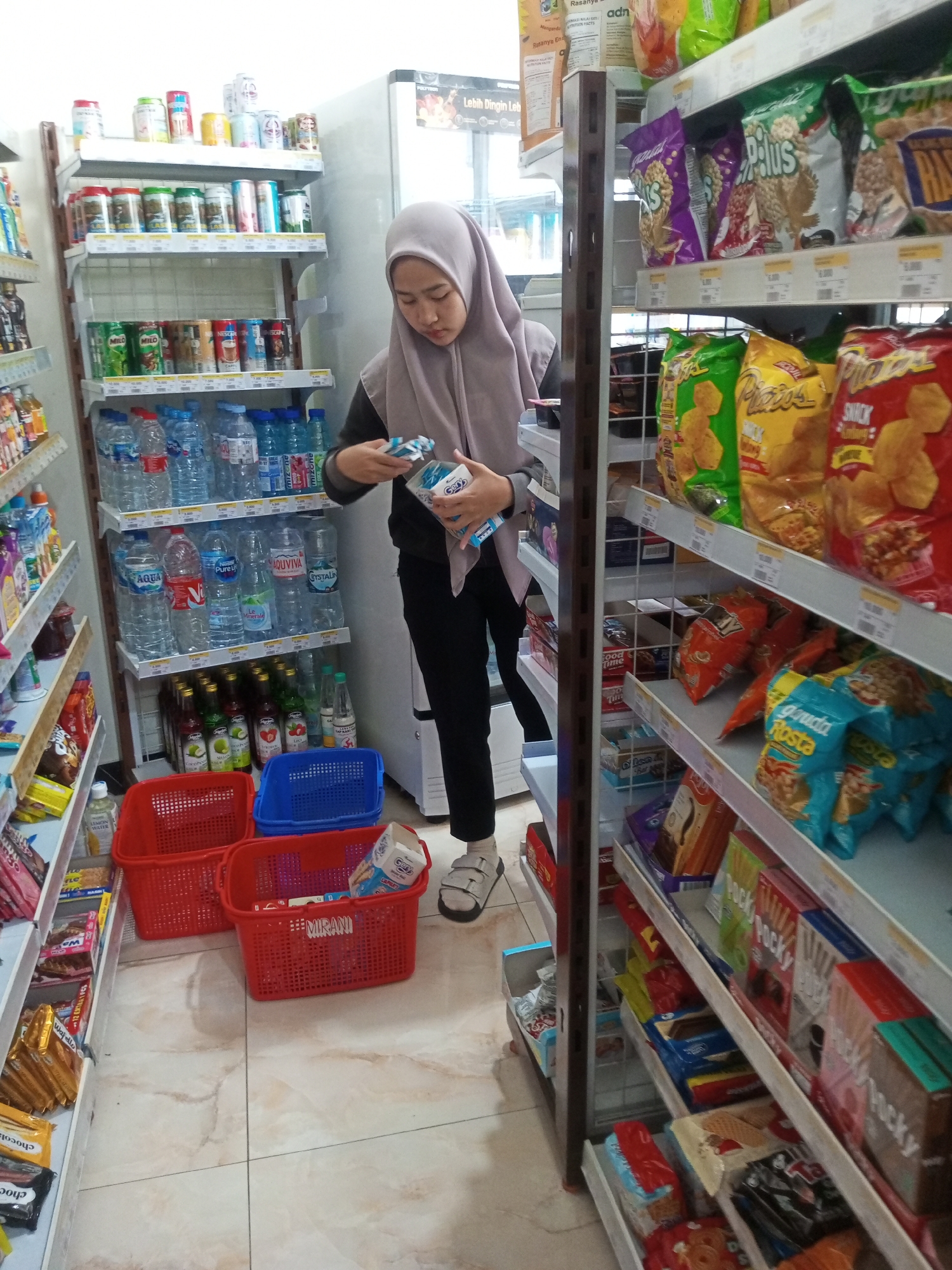 A young woman wearing a light purple hijab and black pants stands in an aisle of a convenience store, examining a packaged product. She is positioned between shelves stocked with various snacks, drinks, and other grocery items. To her left, shelves are filled with bottled water and canned beverages. To her right, shelves display a variety of packaged snacks, including chips and cookies. In the foreground, two red shopping baskets and one blue shopping basket are placed on the floor, with one red basket containing a few packaged items. The store has a tiled floor and a white refrigerator is visible behind the woman. The scene appears to be a typical retail environment within a convenience store.