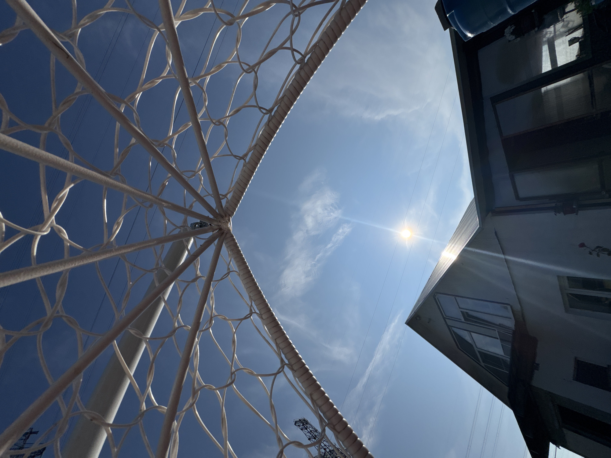 The image depicts a low-angle view from beneath a white, arced structure, possibly a gazebo or sports goal, looking up at a bright blue sky with scattered cirrus clouds. The sun is visible, creating a lens flare effect. In the background, a house with a sloped, dark roof and several windows is partially visible. The house appears to be a residential building located in the city of Chaikovsky, Russia, given the context of the prompt. Power lines are visible crisscrossing the sky, suggesting an urban or suburban environment. The overall scene is an outdoor setting on a clear, sunny day.