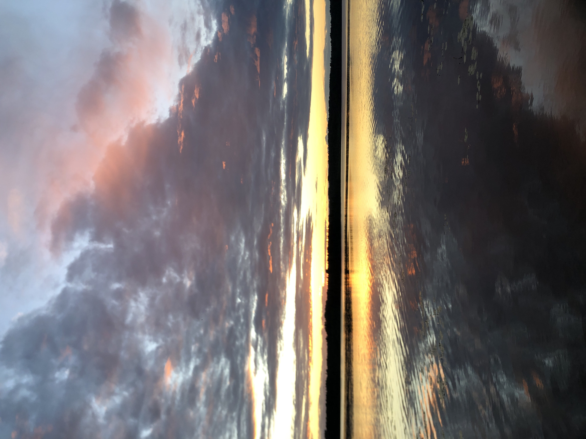 The image captures a tranquil sunset over a body of water, likely a river or lake, in the city of Svirstroy, Russia. The sky is filled with dramatic clouds, tinged with hues of pink, orange, and gray, suggesting the sun is either rising or setting. The water below reflects the sky's colors, creating a mirror-like surface broken only by gentle ripples. A dark silhouette of trees lines the far bank of the water, separating the sky from the water's reflection. The scene is devoid of people or any man-made structures, emphasizing the natural beauty of the landscape.