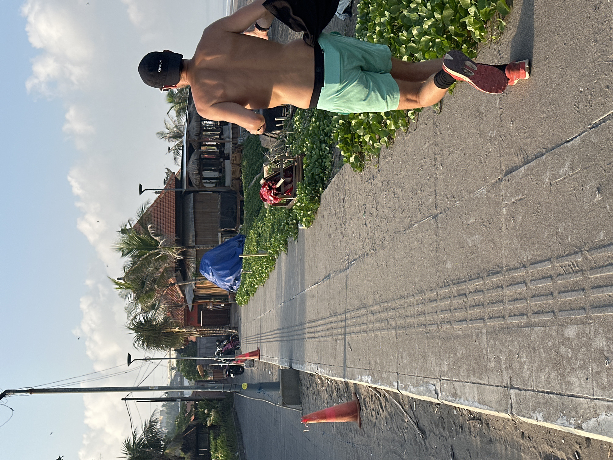A shirtless person with a black baseball cap and light blue shorts is running away from the camera on a paved pathway. They are carrying a black item in their right hand. The pathway is alongside a sandy area with orange traffic cones and some vegetation on the right. In the background, there are buildings with thatched roofs, palm trees, and a clear sky with some clouds. The scene appears to be on a beachside road in North Kuta, Indonesia.