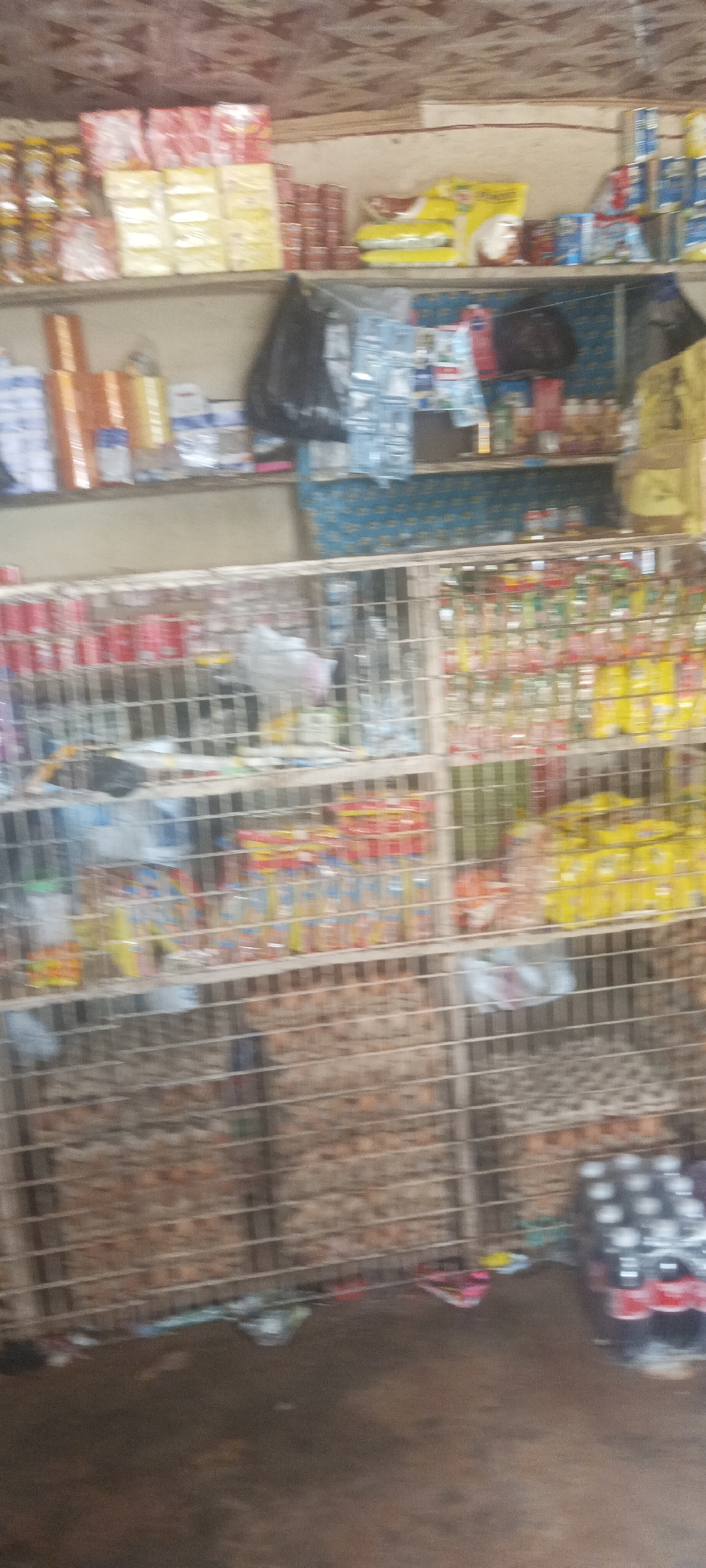 A store in Bokkos, Nigeria. The shelves display a variety of packaged food items and other goods behind a wire mesh. A stack of bottled drinks rests on the floor.