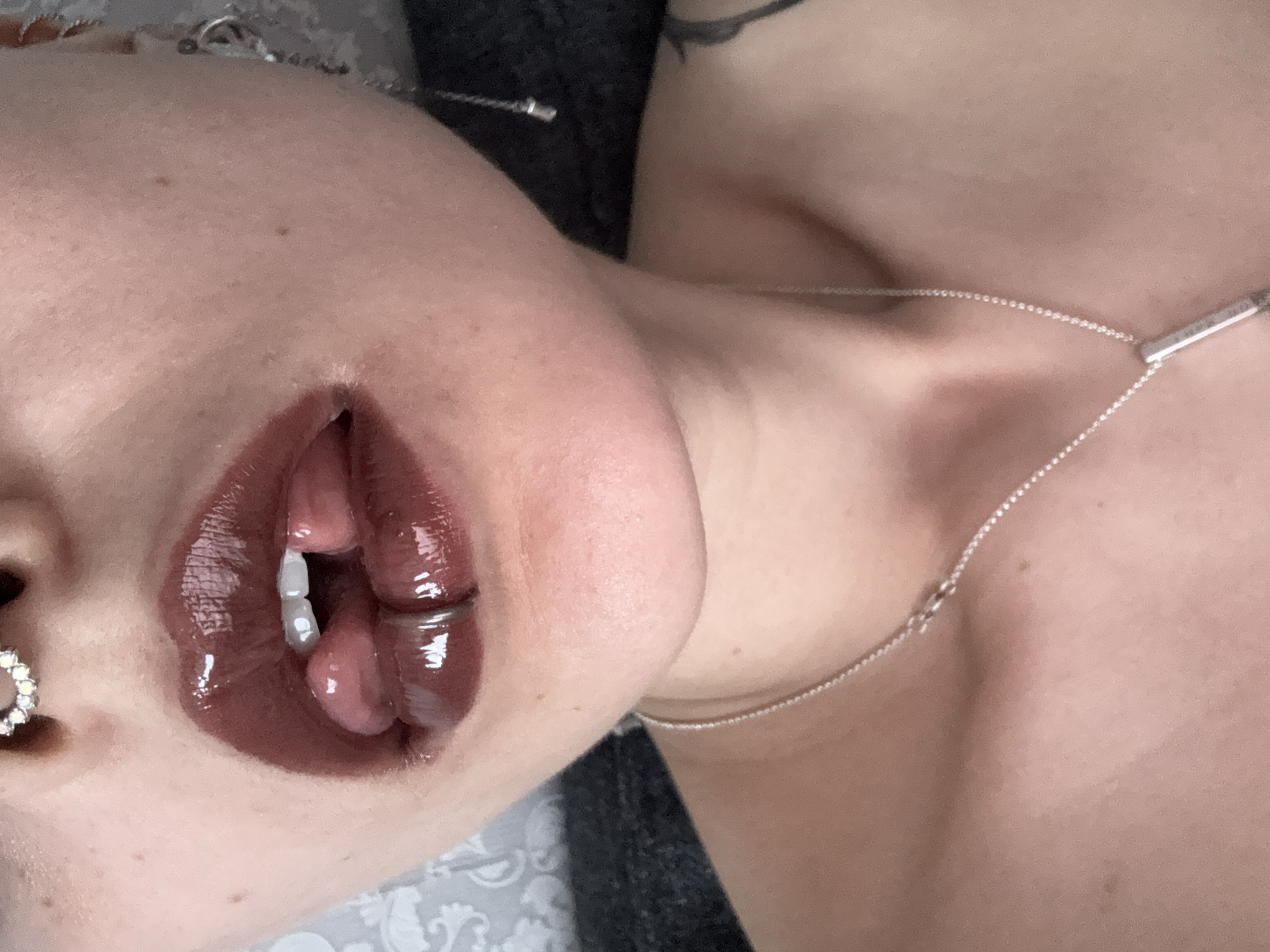 A close-up, angled shot focuses on a person's face, specifically the mouth, with glossy dark lipstick. The person has fair skin and is wearing jewelry: a nose ring and a silver necklace. A glimpse of a tattoo is visible near the shoulder/neck area. The background includes a patterned surface and a dark, undefined area. The location, Saint Petersburg, Russia, cannot be determined from the image.