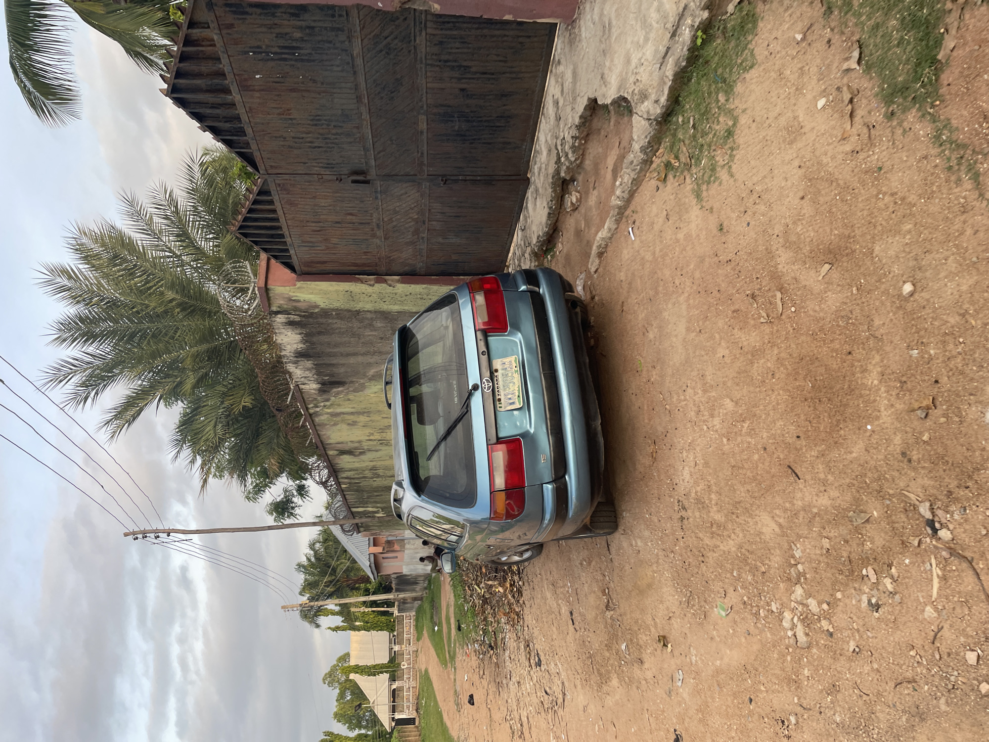 In Jalingo, Nigeria, a turquoise station wagon is parked on a dirt road in front of a large, corrugated metal gate. The car, a Toyota, has a license plate that reads "WKK 585AA". The road is covered in red dirt and scattered debris, including leaves and small stones. To the left of the car, a low, green wall runs along the property line, topped with barbed wire. Beyond the wall, palm trees and other foliage are visible. Two individuals are seated near the wall, to the left of the car, appearing to be resting or observing. The sky above is overcast with clouds.