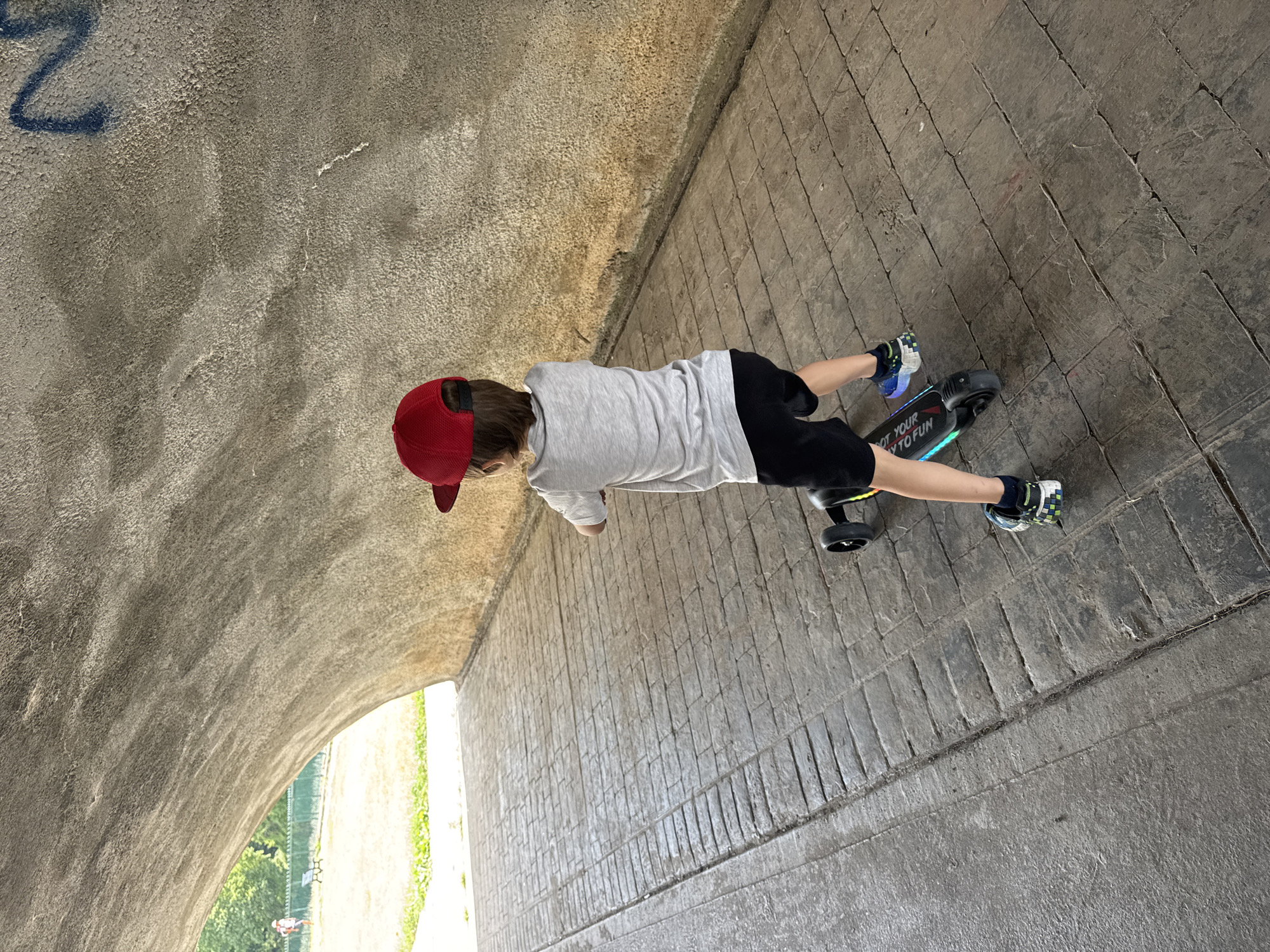A young child wearing a red baseball cap and a grey t-shirt with black shorts rides an electric scooter through a textured concrete tunnel. The scooter has the words "NOT YOUR" and "TO FUN" visible on its deck, along with colorful LED lights. The child is facing away from the camera, looking towards the tunnel exit, where daylight illuminates a grassy area with a fence and trees. The tunnel's interior walls are rough concrete, and the ground is paved with rectangular stone tiles. Graffiti is visible on the tunnel ceiling.