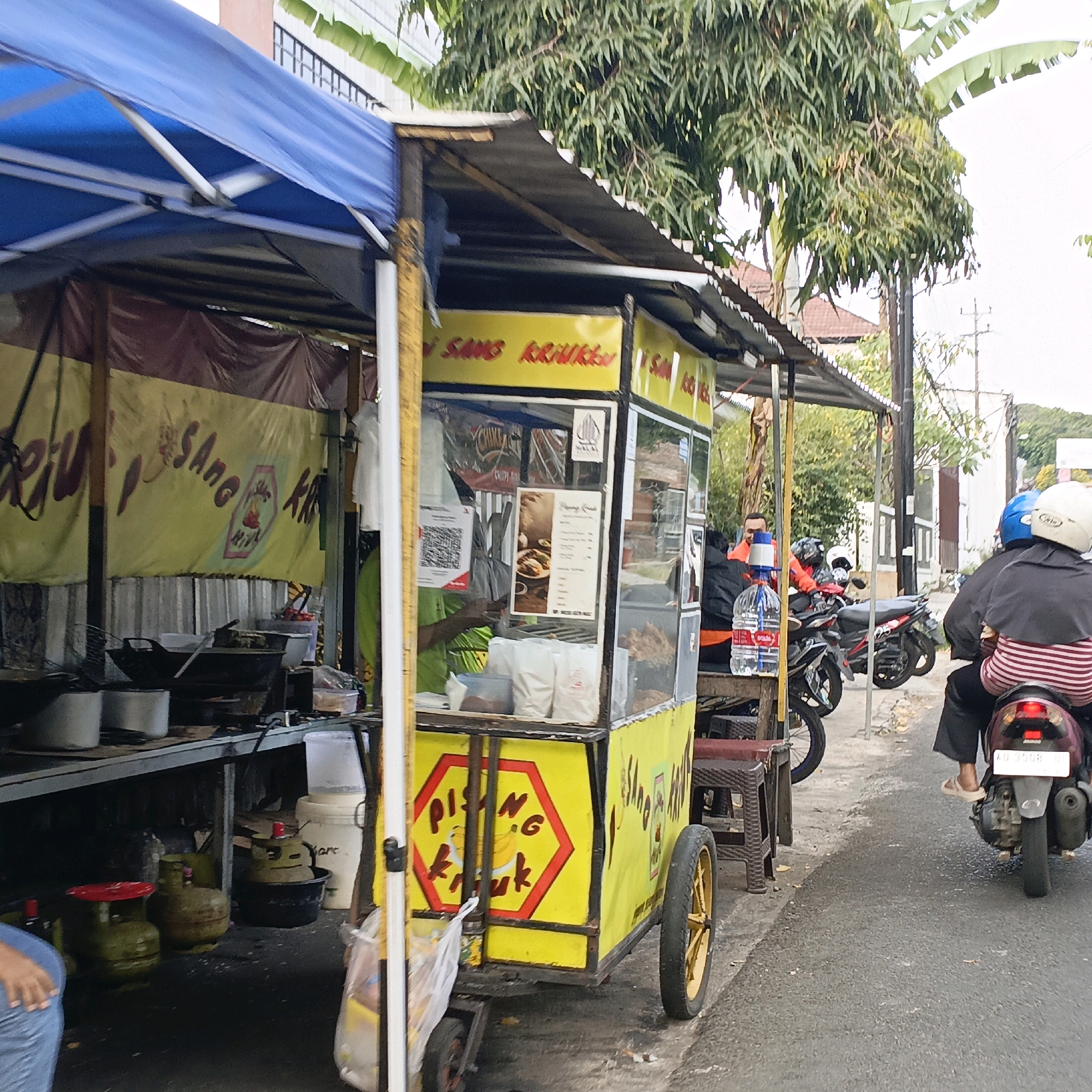The image depicts a street scene in Salatiga, Indonesia, focusing on a food stall selling fried bananas. The stall is characterized by a bright yellow cart with a blue tarp overhead. The cart is laden with cooking equipment, including large woks and gas cylinders, suggesting active food preparation. A banner with Indonesian writing, likely advertising the food, is visible.
Several people are present in the scene. Two individuals are on a motorcycle, one driving and one a passenger wearing a striped shirt and a black headscarf, indicating they are likely customers. Another person, wearing an orange shirt and a blue head covering, is seated near the stall, possibly a vendor or another customer. In the background, more motorcycles are parked, and buildings line the street. The overall environment appears to be a bustling urban setting, typical of a street-side market or food vendor area. The lighting suggests it is daytime.