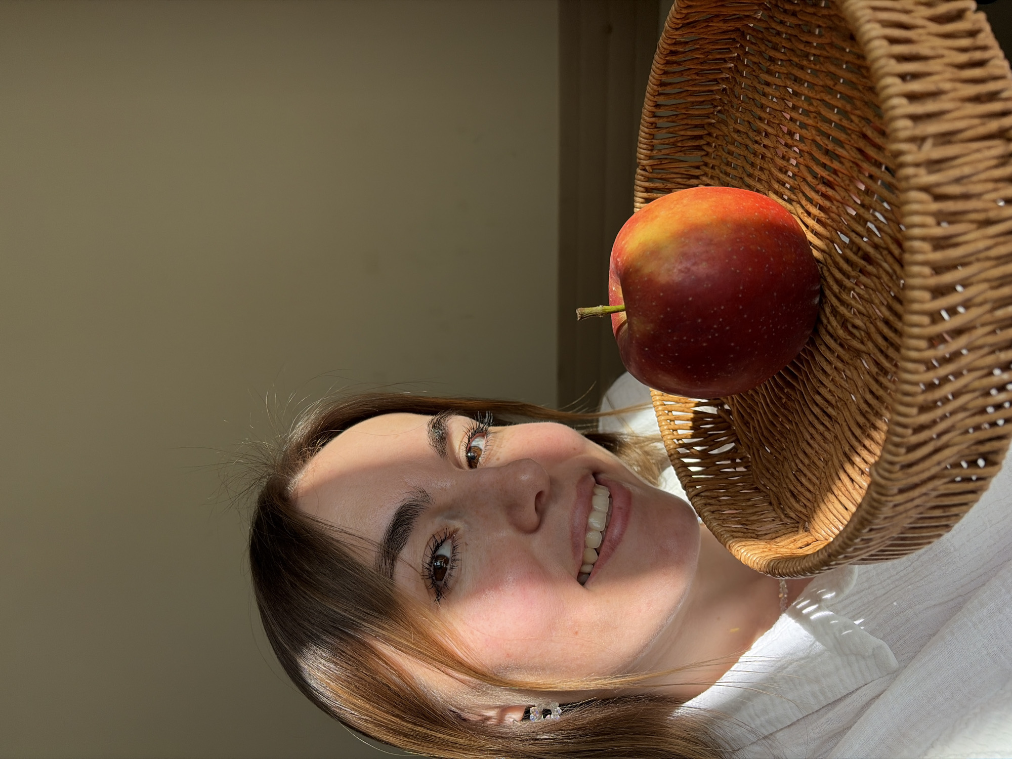 A person, presumably female based on the presence of long hair and facial features, is holding a wicker basket containing a red apple with a green stem. The background appears to be an interior wall. There is no information to determine if the location is in the city of Izhevsk, Russia, as there are no identifiable landmarks or features to place it.