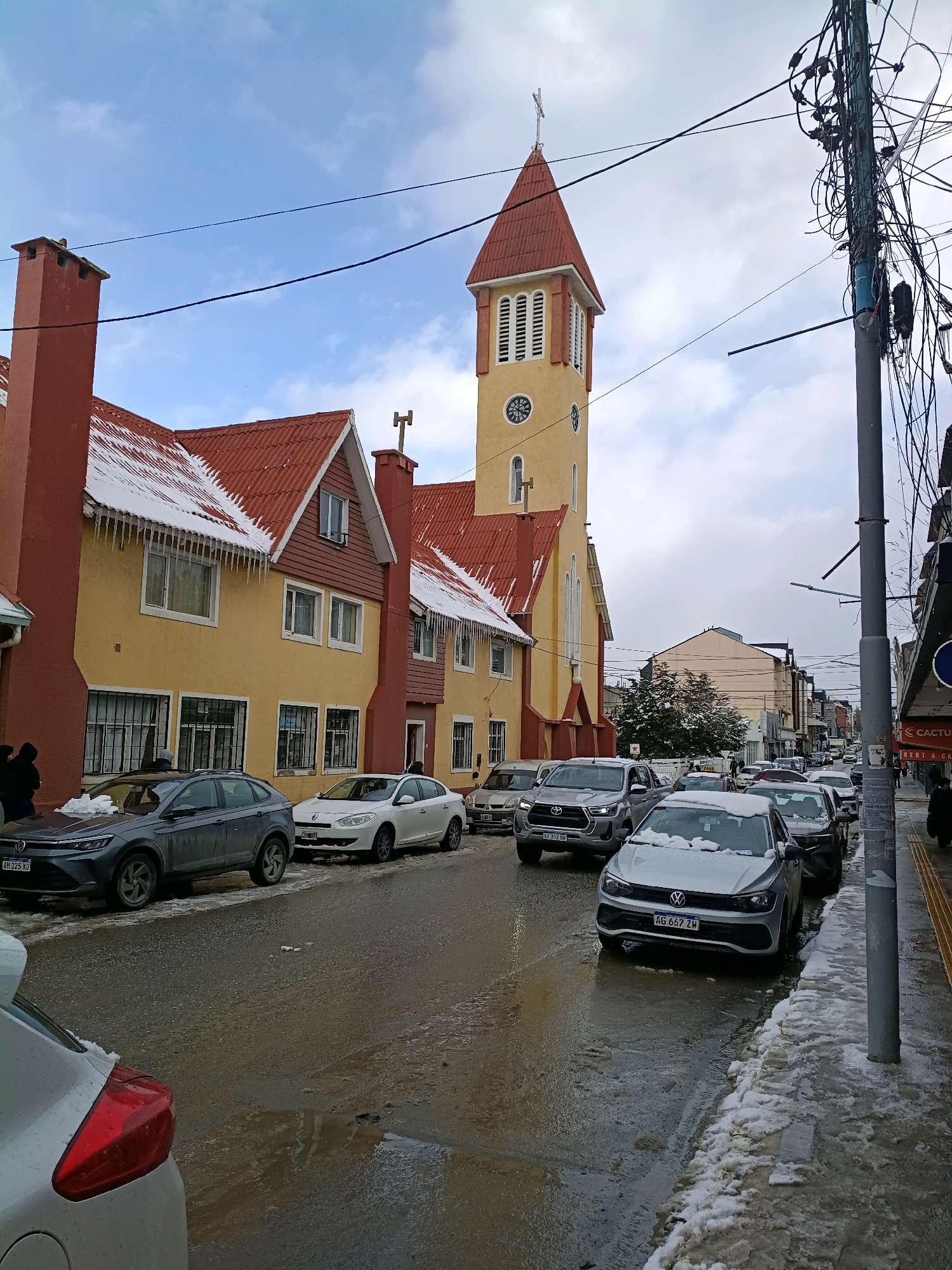 The image depicts a street scene in Ushuaia, Argentina, likely during winter, given the presence of snow and icicles on the rooftops. A prominent yellow church with a red-tiled steeple stands on the left side of the street, adjacent to a building with a red roof and chimneys. The street is lined with parked cars, some of which are covered in snow. The road surface appears wet and partially covered with snow and slush. Electrical poles with a dense network of wires are visible on the right side of the image, indicating urban infrastructure. A few pedestrians are visible on the sidewalk and near the buildings. The sky is overcast with some blue patches. The overall impression is of a cold, urban environment.