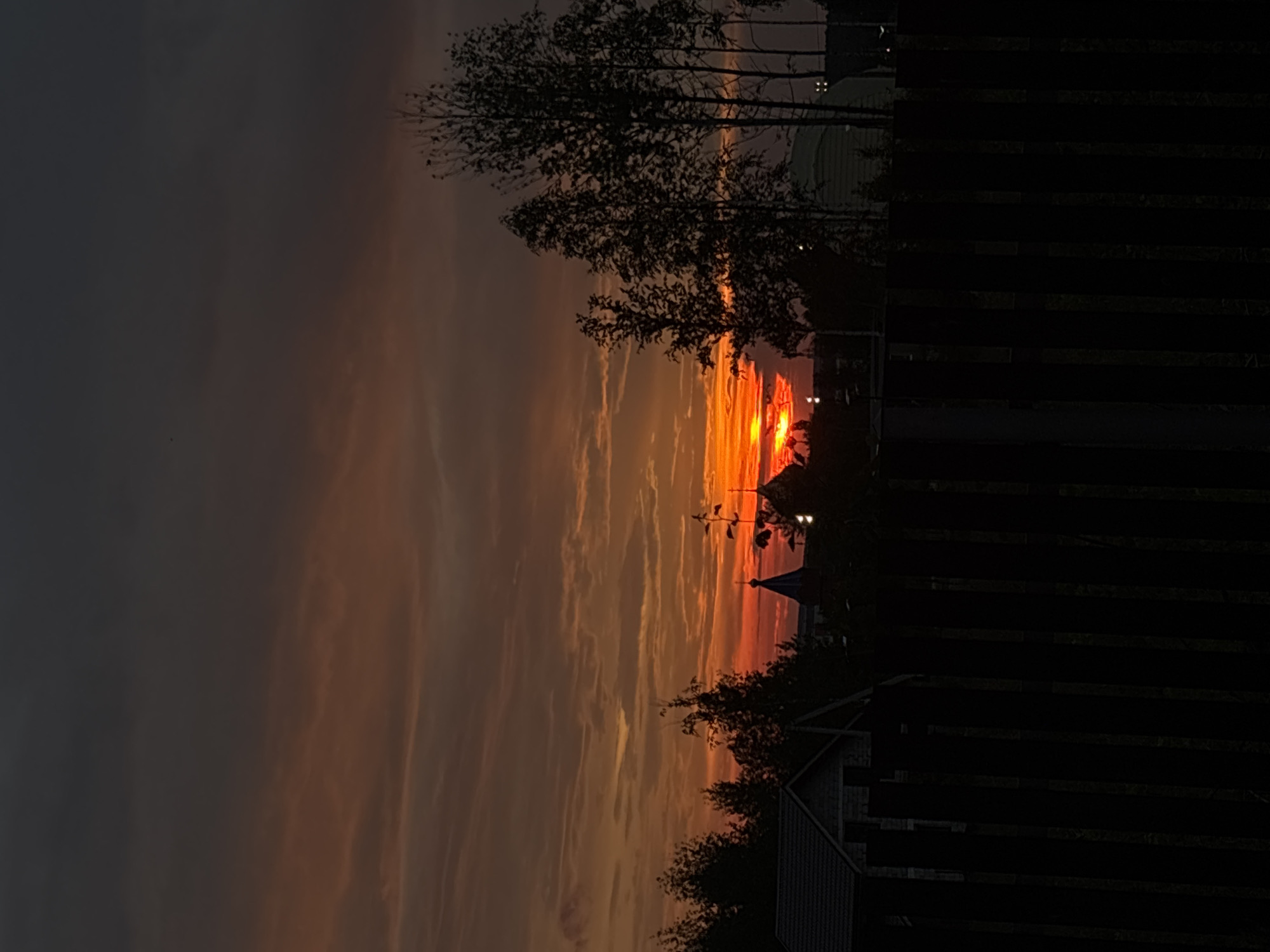 The image depicts a sunset with a vibrant orange and red sky over Gubakha, Russia. In the foreground, a dark wooden fence runs horizontally across the frame. Behind the fence, silhouetted trees and buildings are visible against the dramatic sky. Among the structures, two domed buildings, likely churches, stand out. The overall scene is one of natural beauty at dusk, with a touch of local architecture. There are no people visible in the image.