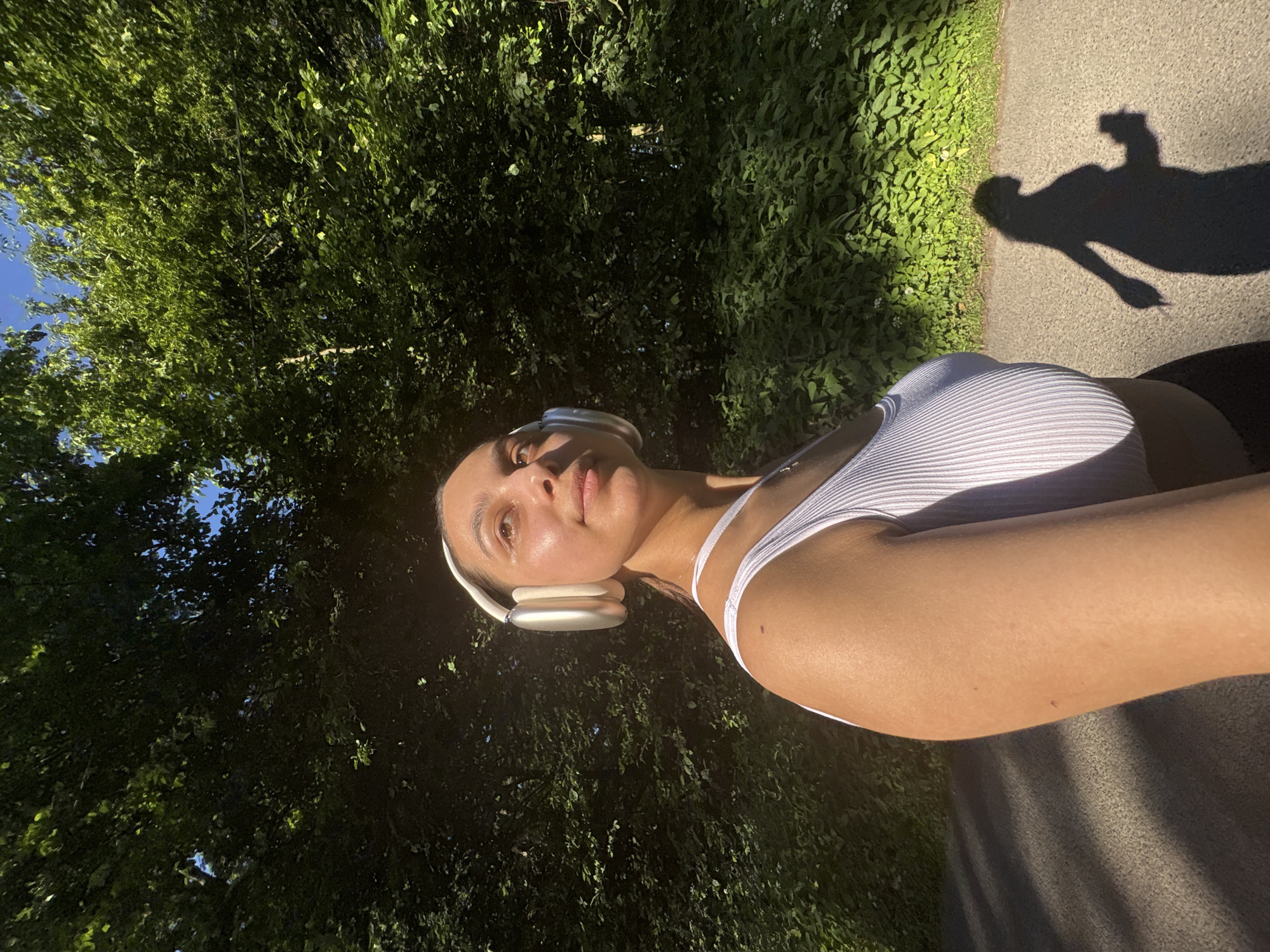 A woman in a white tank top and white headphones is walking on a paved path, likely in a park. Her shadow is visible on the path. The path is lined with green vegetation on both sides. The location appears to be outdoors with trees visible. It is daytime, judging by the bright sunlight and shadows. Without further information, it is difficult to pinpoint the exact location in Mönchengladbach.