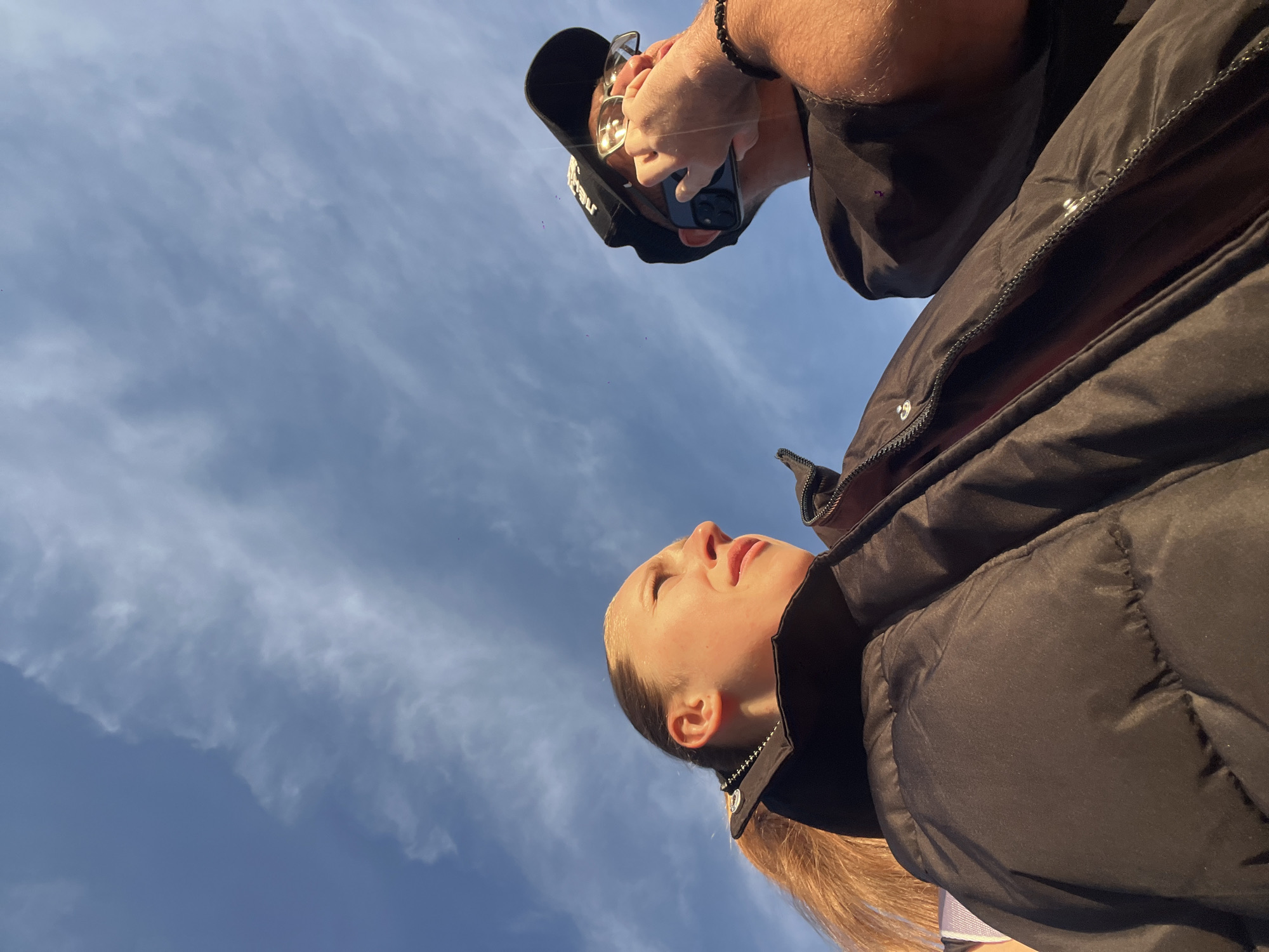 The image shows two people from a low angle, with a blue sky and scattered clouds in the background. A woman with blonde hair tied in a ponytail is in the foreground, looking upwards. She is wearing a black puffer jacket. Above her is a man wearing a black baseball cap, sunglasses, a beaded bracelet, and a black mask. He is holding a cell phone up in front of his face. The location is unknown.
